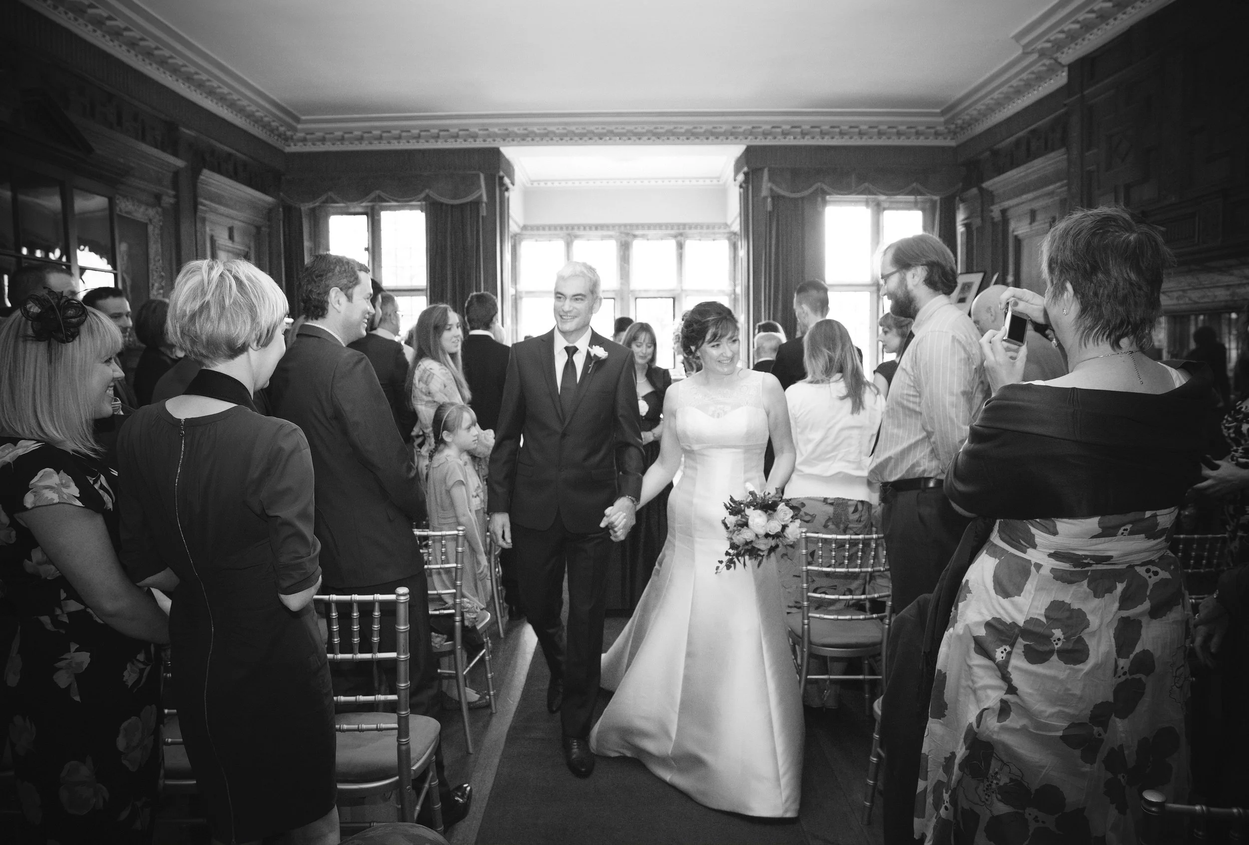 A wedding ceremony with a bride and groom holding hands, surrounded by guests. The bride is wearing a white wedding dress and holding a bouquet of flowers. The groom is dressed in a black suit with a boutonniere. Guests are smiling and some are takin