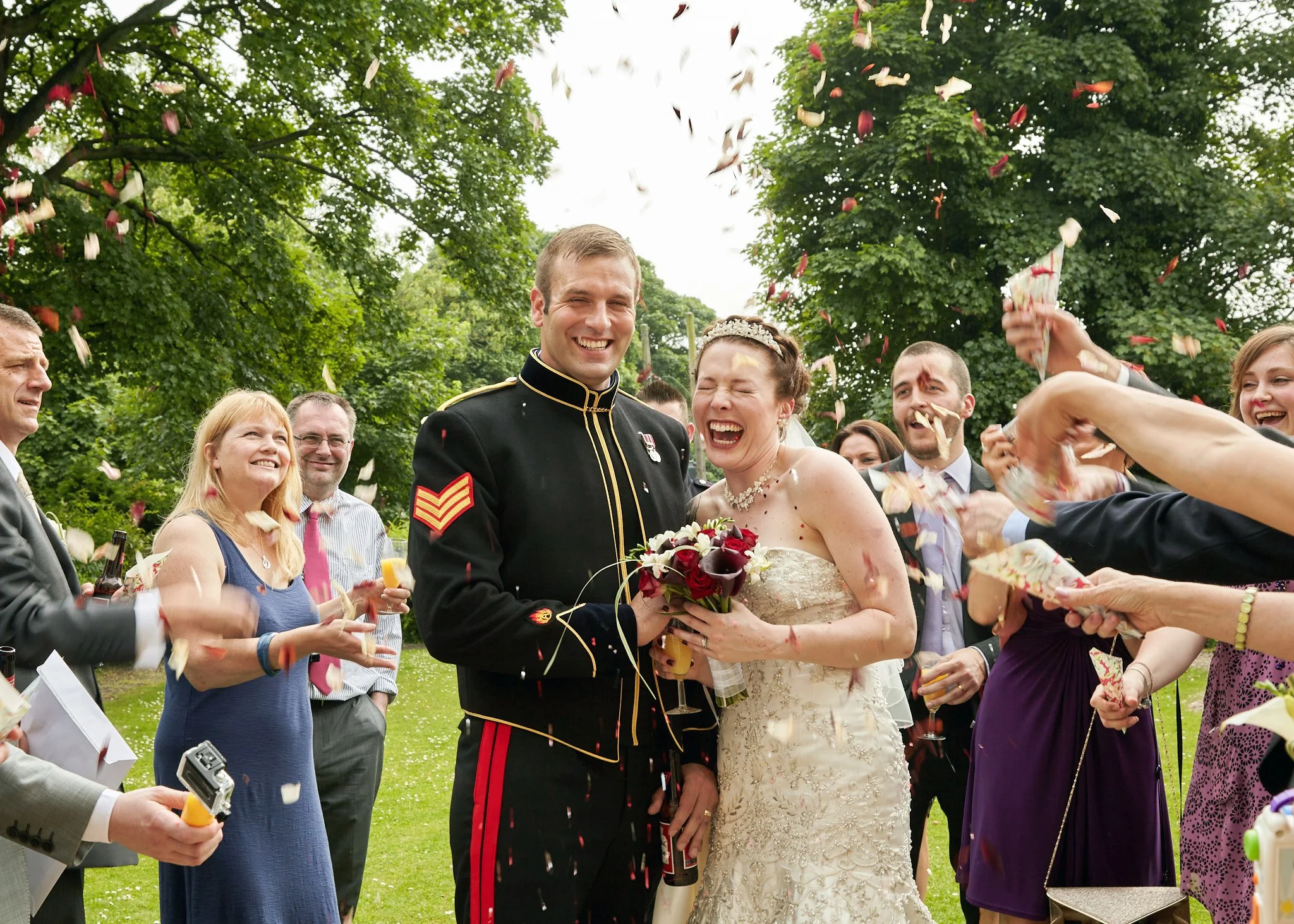 Bride and groom celebrating wedding outdoors with friends and family, confetti falling, trees in background.