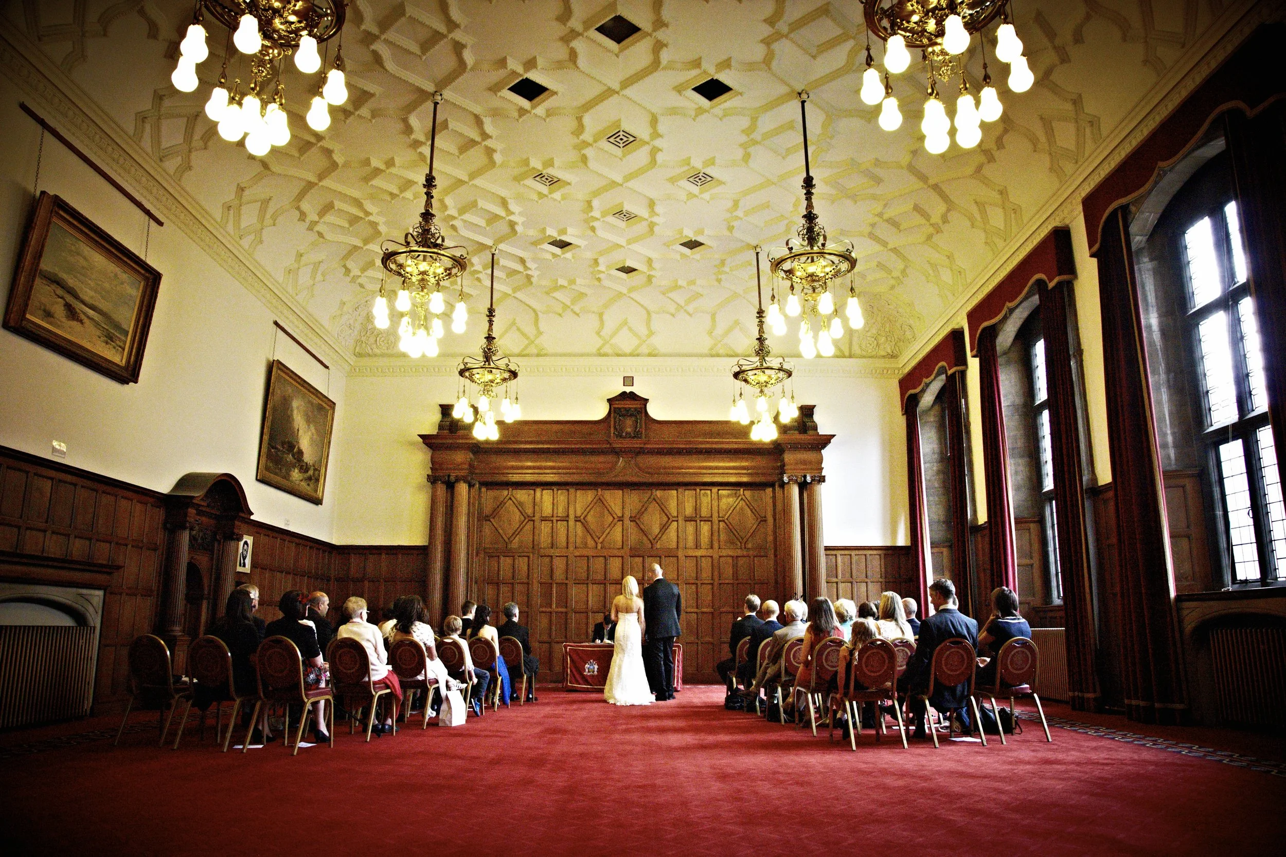 A wedding ceremony taking place in an ornate, historic room with high decorative ceiling, chandeliers, large windows, and wood paneled walls. The bride and groom are standing in front of an officiant, facing each other, with guests seated in rows of chairs watching the ceremony.