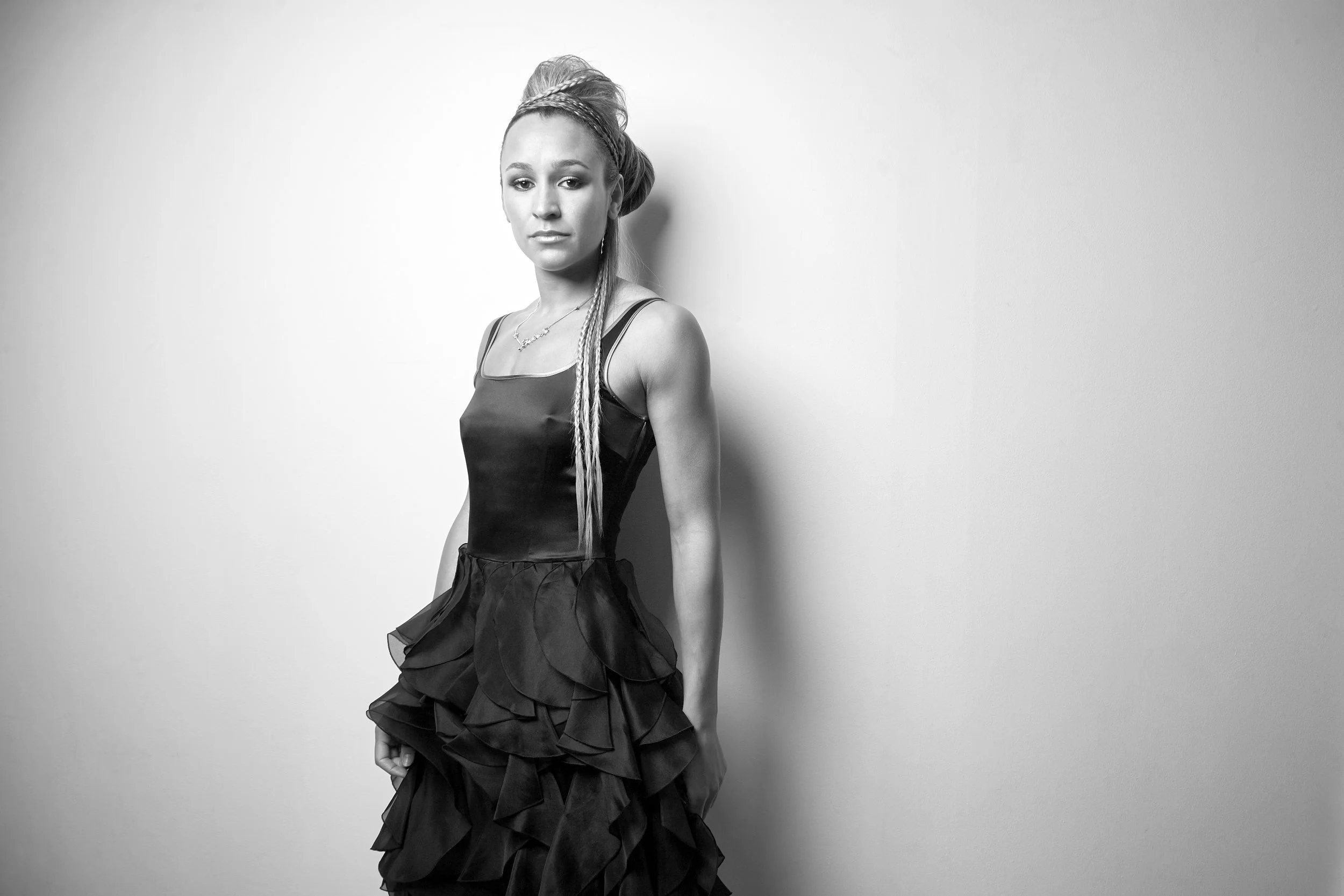 Black and white photograph of a woman with styled hair and long, braided extensions, wearing a sleeveless top and a layered, ruffled skirt, standing against a plain wall.