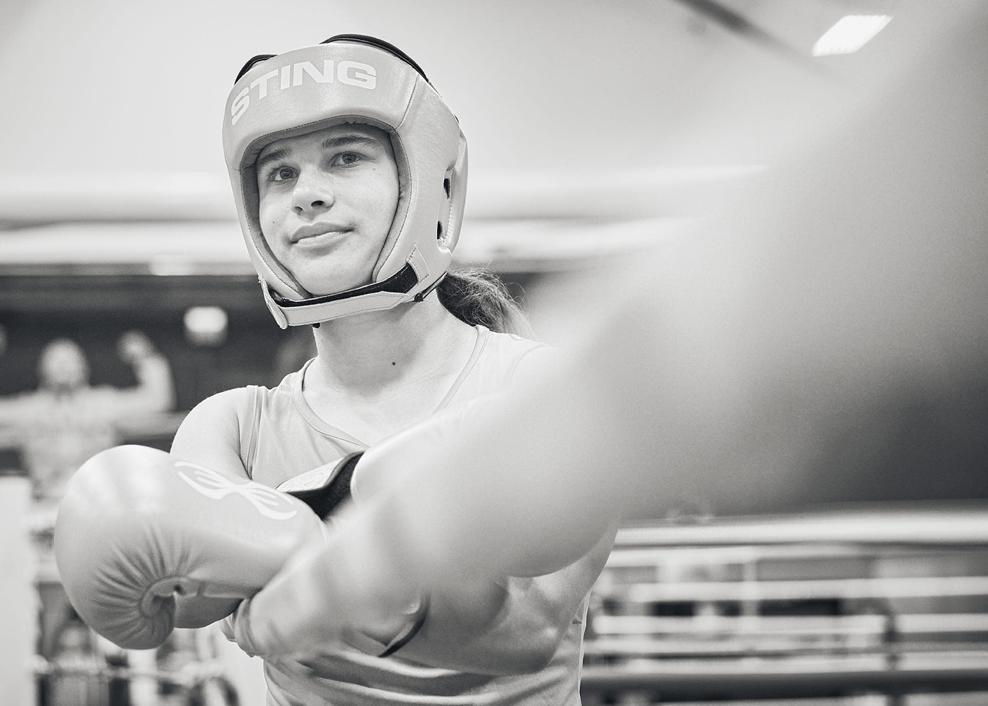 A young woman wearing boxing gear, including a head guard and gloves, practicing or sparring in a gym.