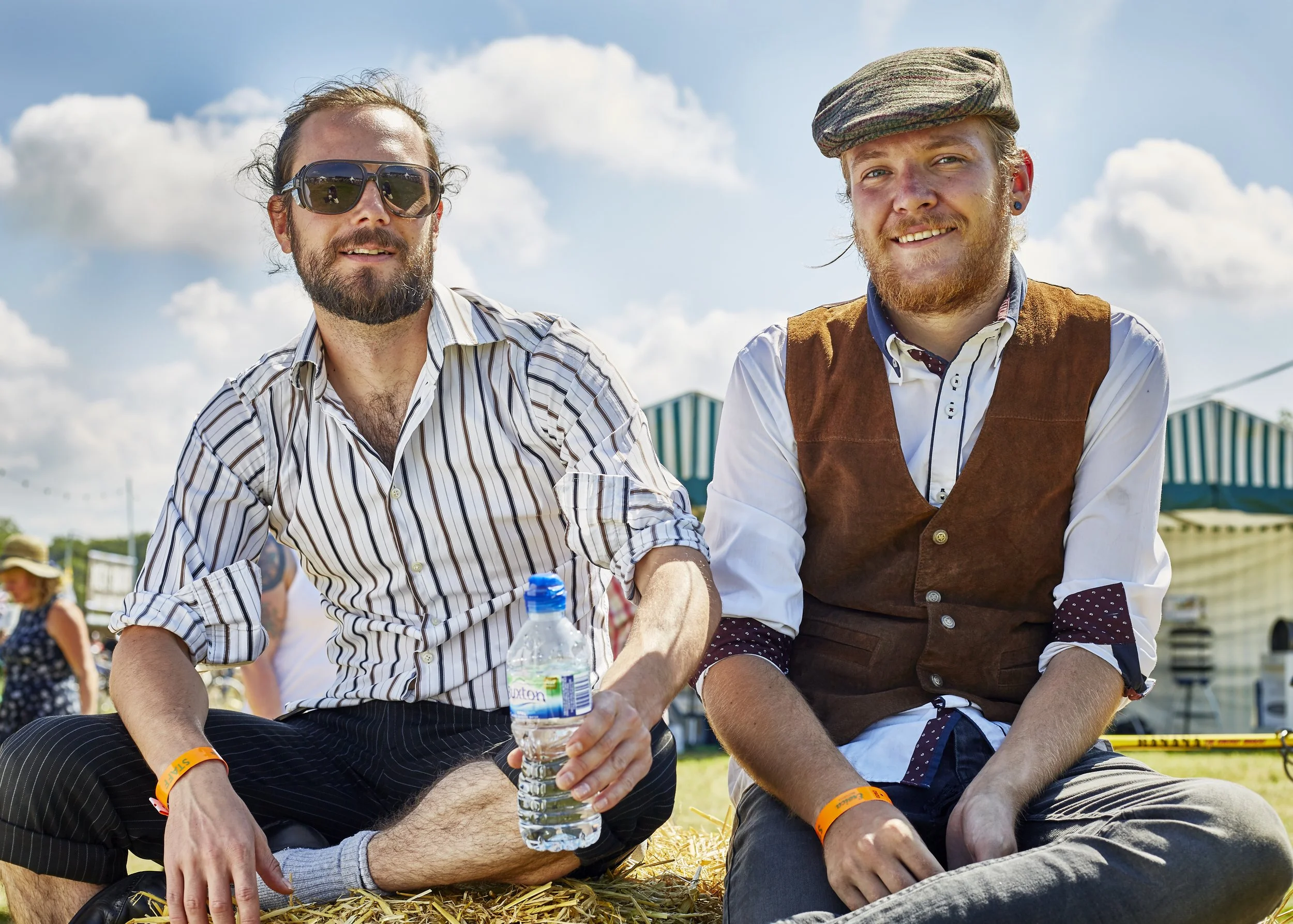 Two young men sitting outdoors on hay bales at a festival or fair on a sunny day, with blue sky and clouds in the background. One wearing sunglasses and a striped shirt, holding water bottle; the other wearing a flat cap, vest, and polka-dot shirt.