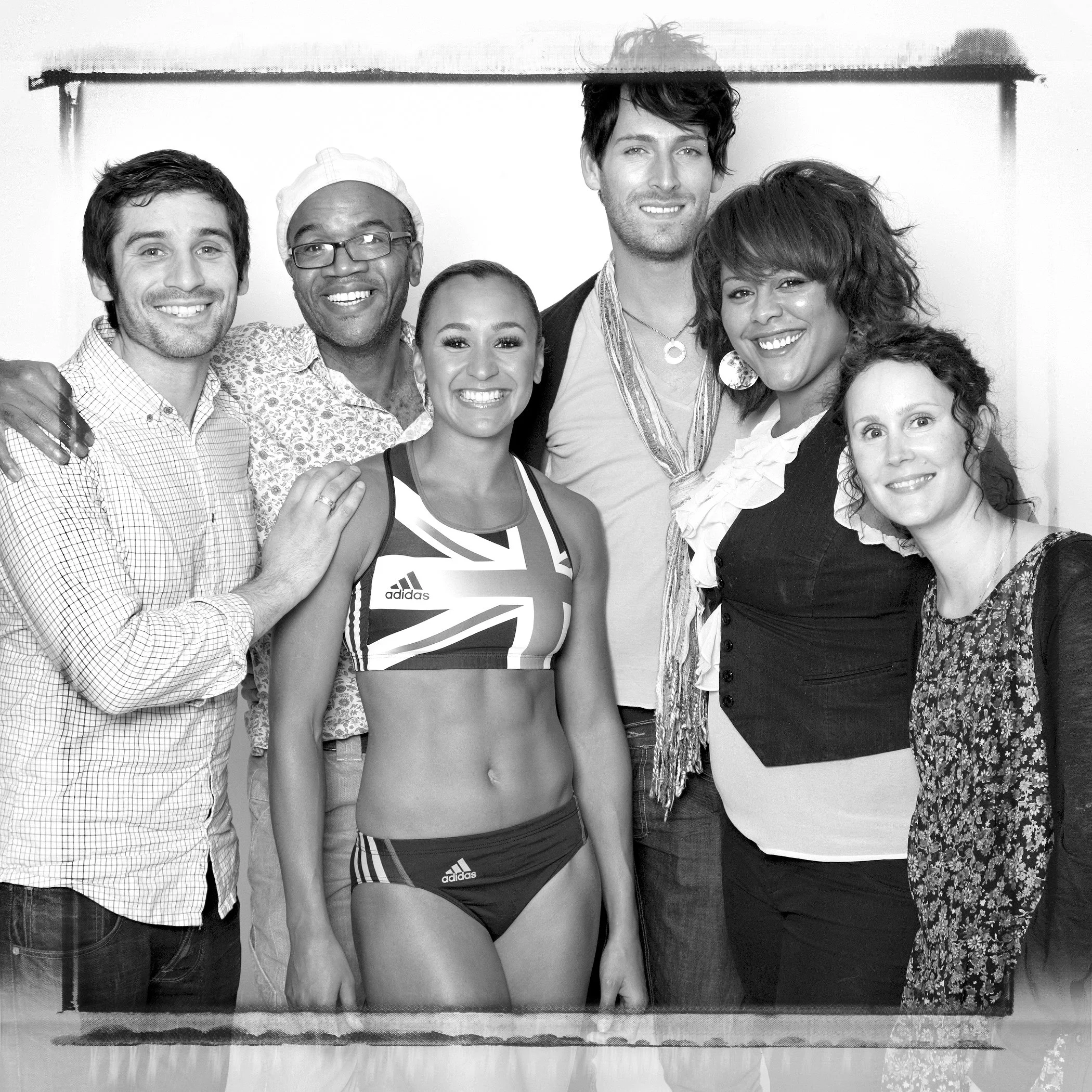 Black and white photo of a diverse group of six smiling people, including men and women of different ethnicities, standing close together in front of a plain background.