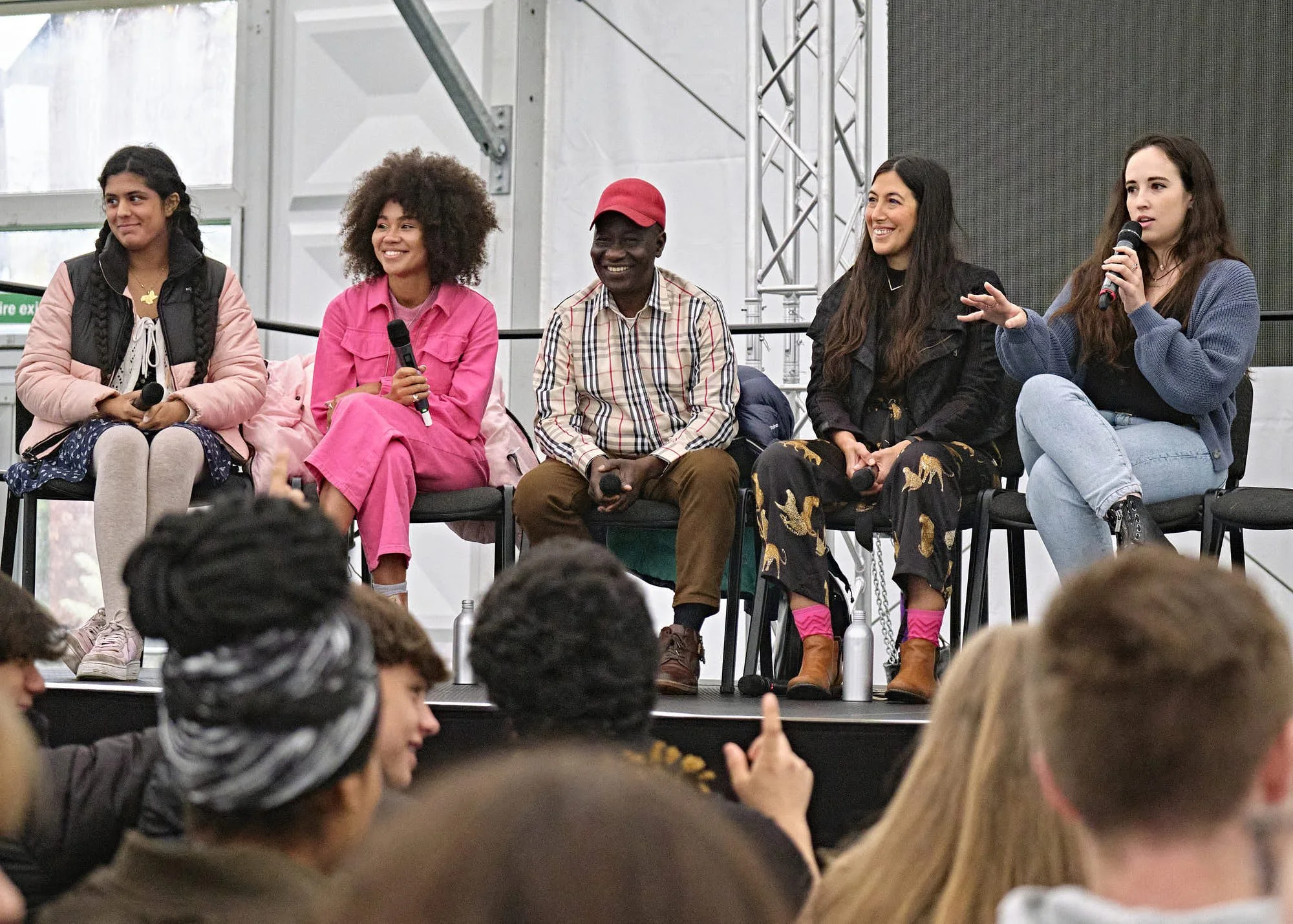 A diverse group of five people sitting on a stage during a panel discussion at an event with an audience in front. Four women and one man are holding microphones and smiling, with one woman speaking into a microphone.