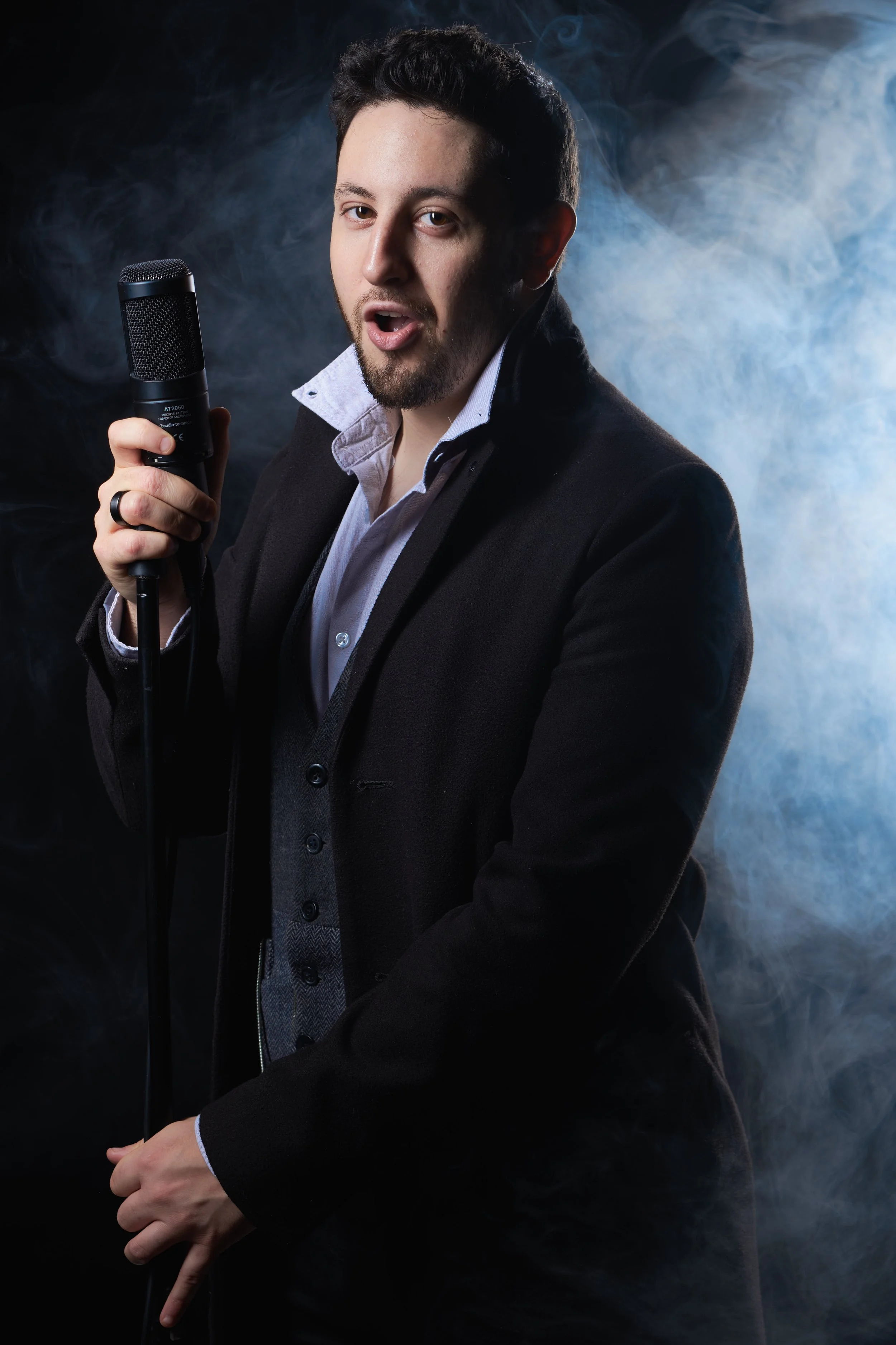 A man with dark, curly hair and a beard holding a microphone, wearing a black blazer and a white shirt, standing against a smoky, dark background.