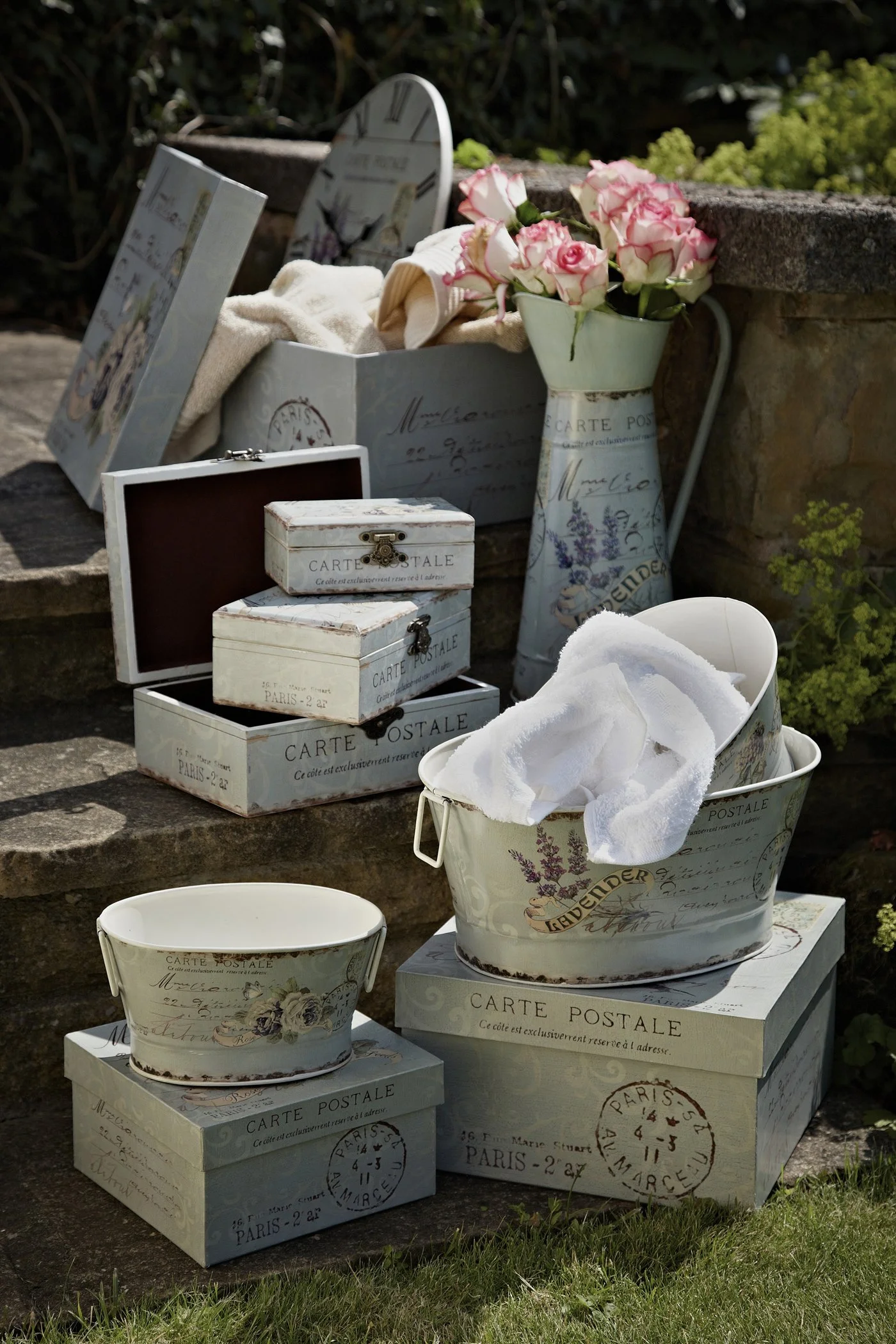 Decorative vintage-style postal-themed boxes and a vase with pink and white roses outdoors on stone steps, with a white cloth and greenery in the background.