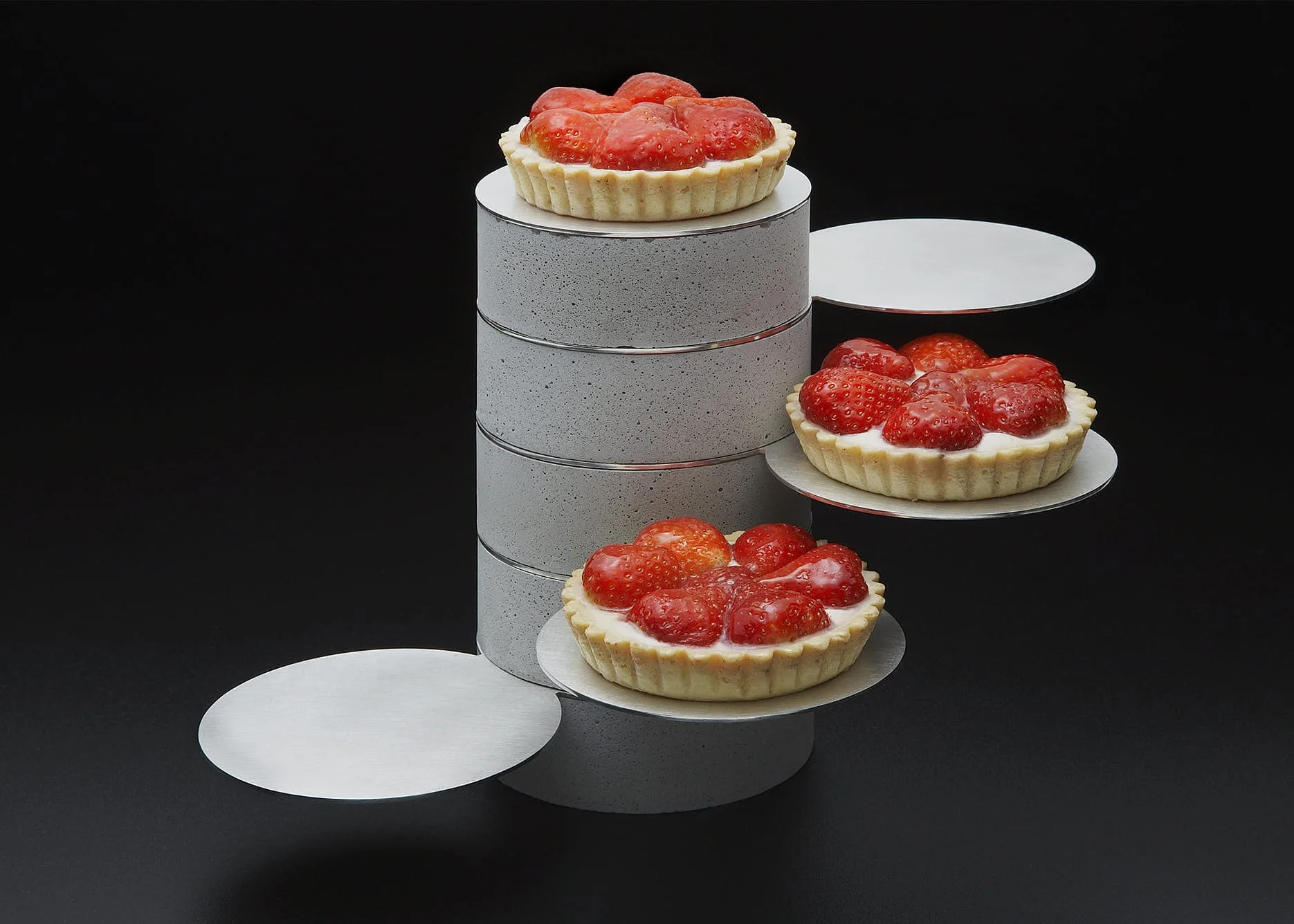 Several strawberry tart cakes placed on white circular platforms against a black background, with some cakes on graduate rings.