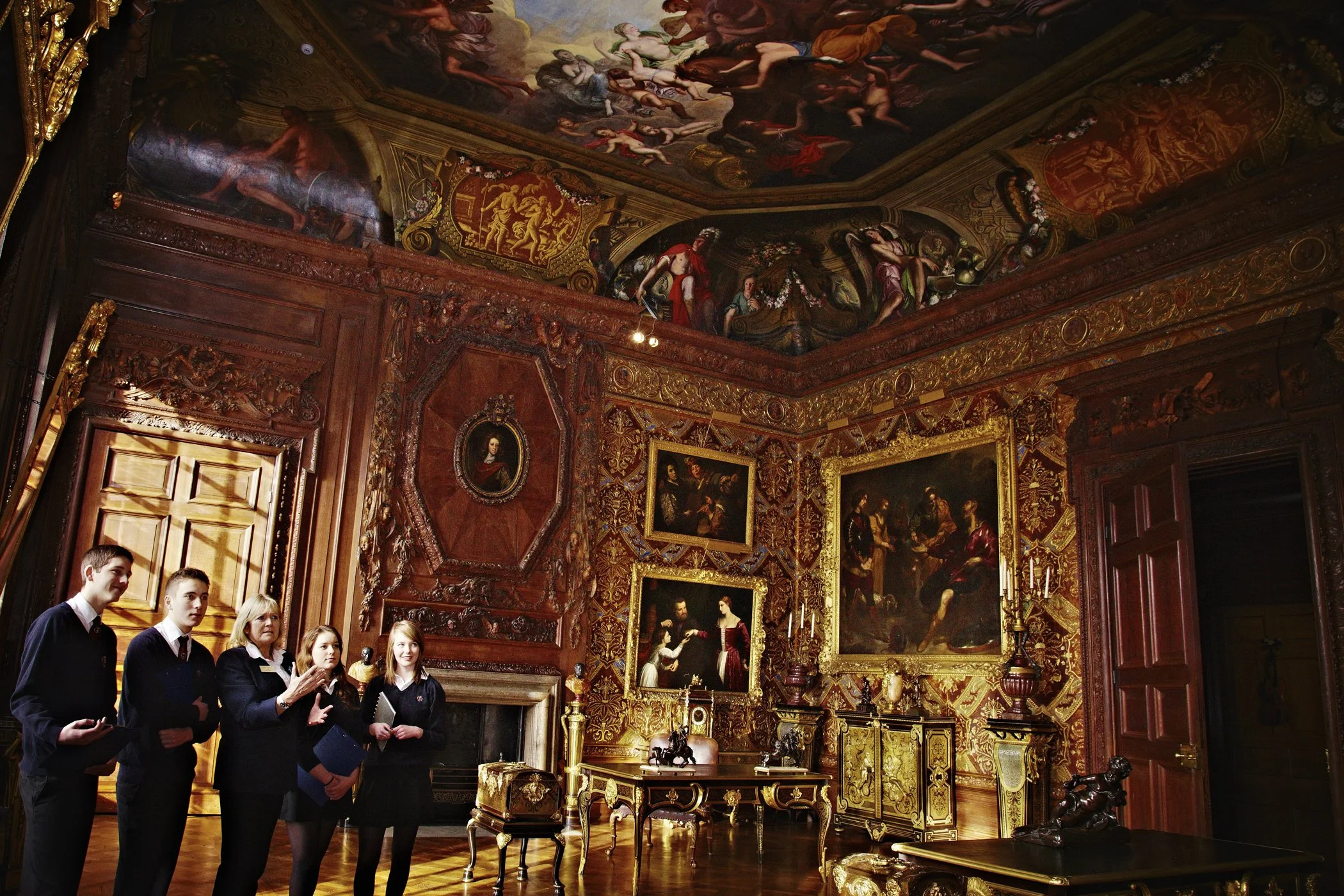 A group of young students on a tour inside a lavishly decorated historical room, featuring ornate woodwork, gold accents, and large classical paintings on the walls and ceiling.
