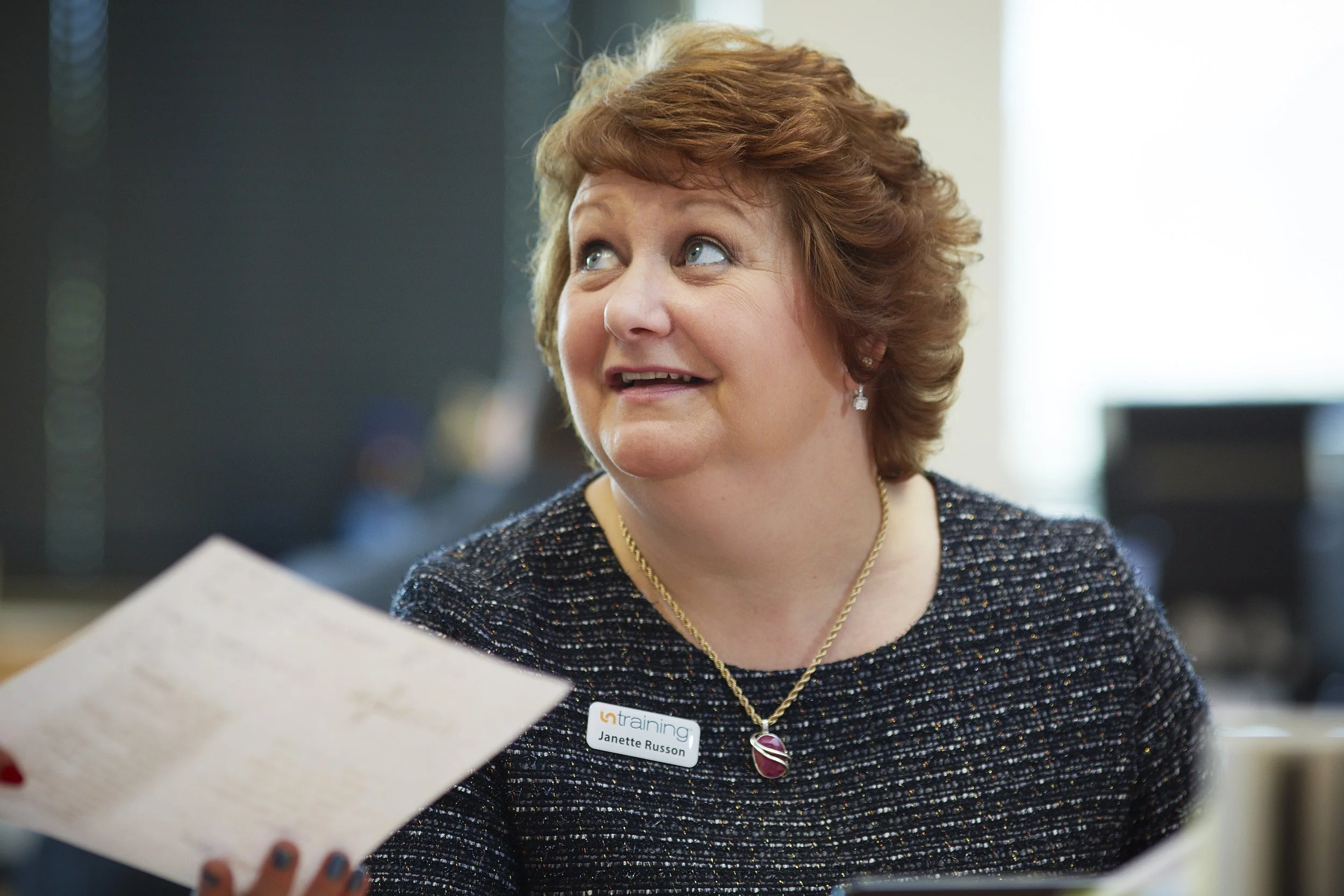 A woman with short red hair wearing a black sparkle dress, a gold necklace, earrings, and a name tag that says 'Janette Russon', looking up and smiling. She is inside a room with a blurred background.