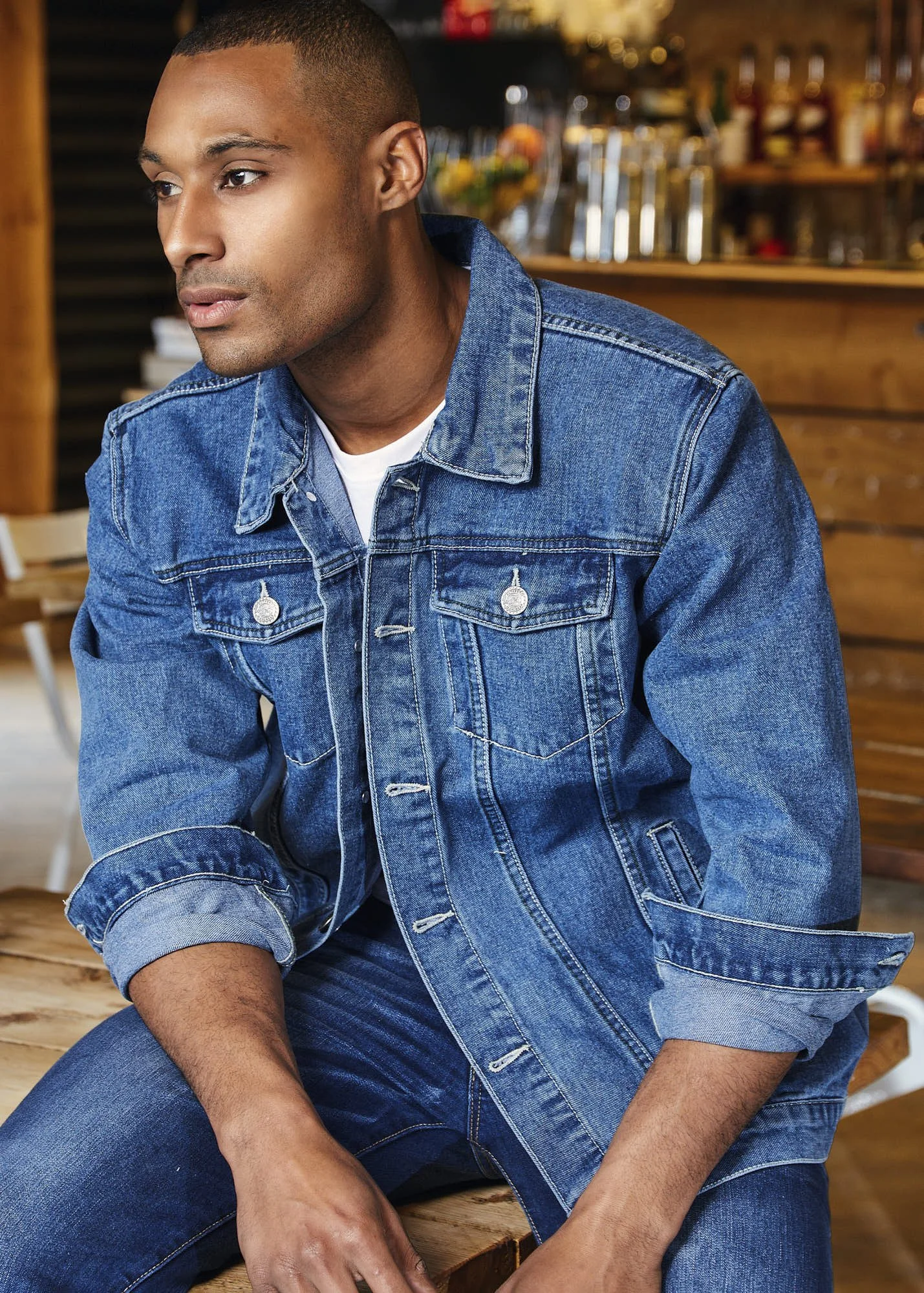 A young man with short black hair and medium skin tone, wearing a denim jacket with the sleeves rolled up, sitting at a wooden table in a cozy cafe or bar with wooden shelves and bottles in the background, looking to the side with a neutral expressio