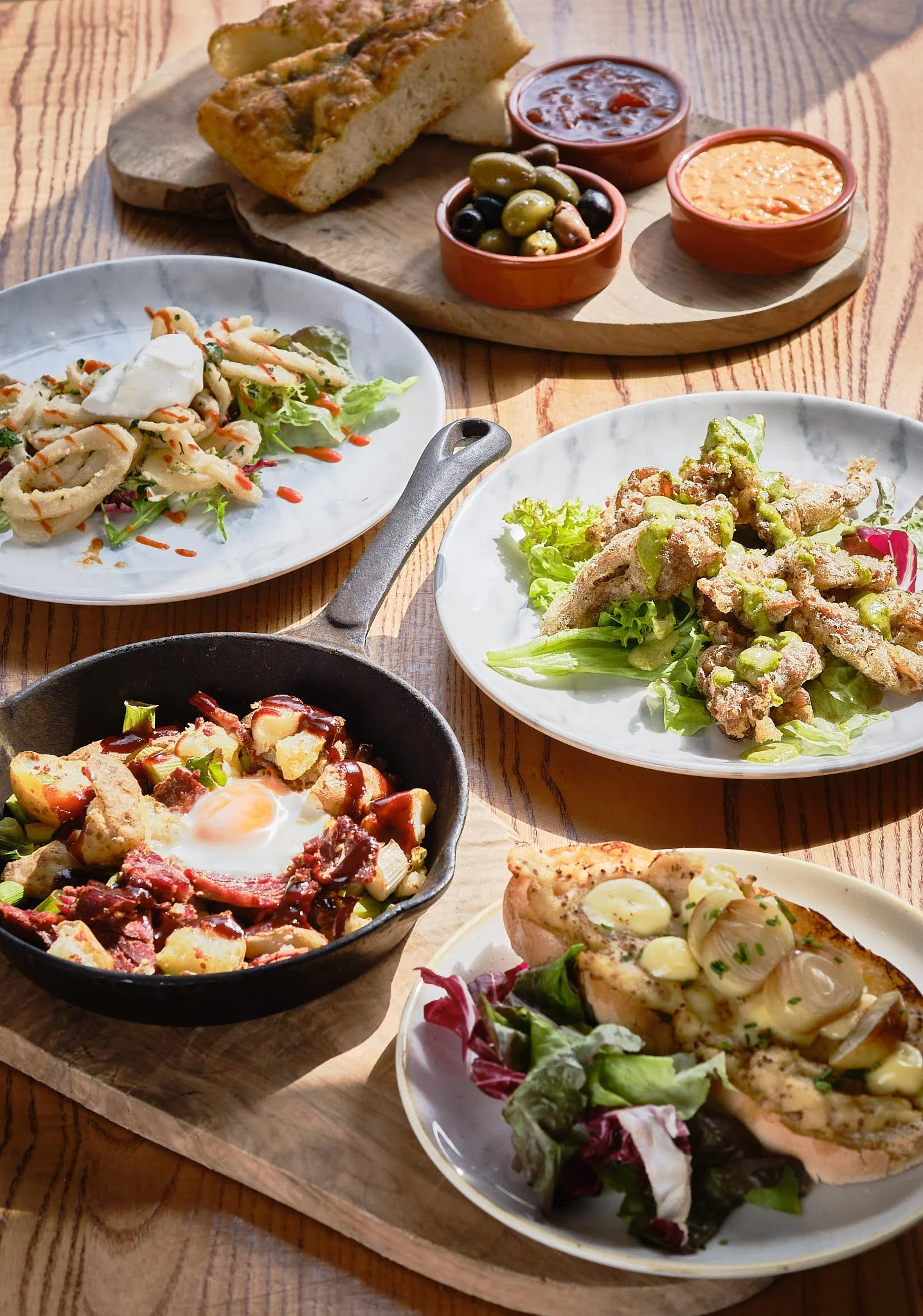 Assorted appetizers and entrees on wooden platters and plates, including a slice of bread topped with cheese, a small bowl of olives, bowls of chili or stew, fried chicken with green sauce, salad with egg and bacon, and a garlic bread slice on a tabl