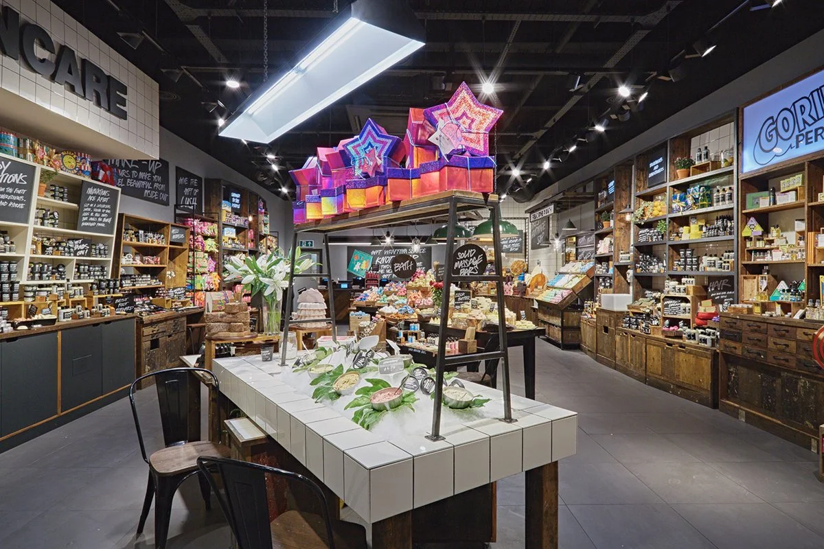 Interior of a gift shop with shelves stocked with various products, a display table with soaps and plants, and decorative light fixtures hanging from the ceiling.