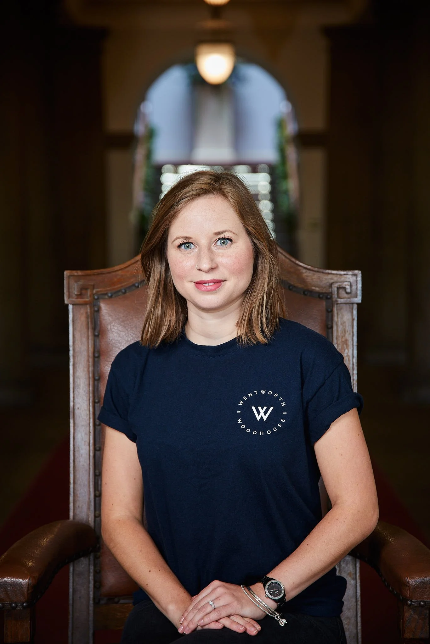 A young woman with light brown hair and blue eyes sitting in a wooden armchair indoors, wearing a navy blue T-shirt with white text and a logo, with a large window in the background.