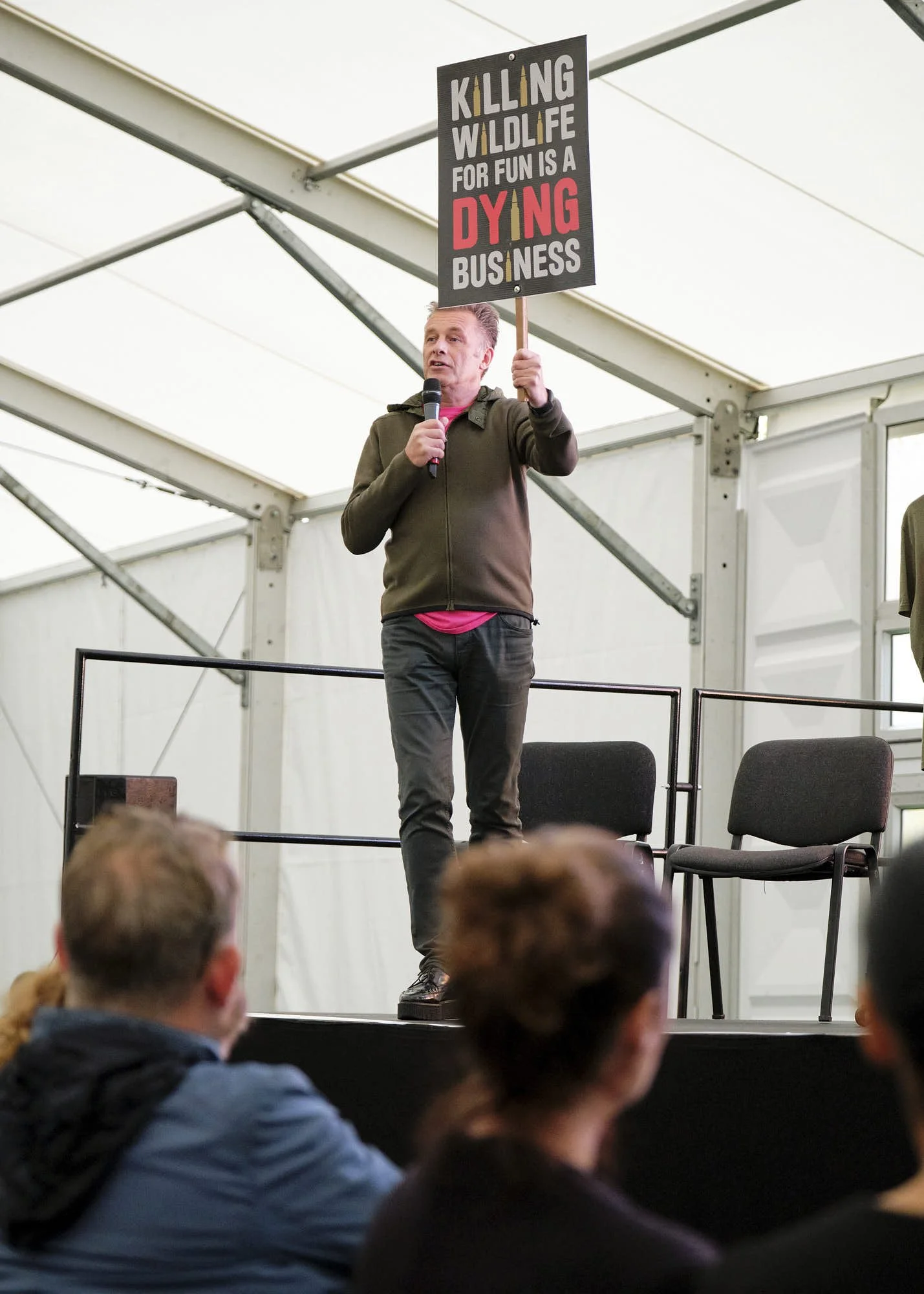 A man holding a microphone and protest sign that reads 'Killing wildlife for fun is a dying business' while speaking at an indoor event with an audience.