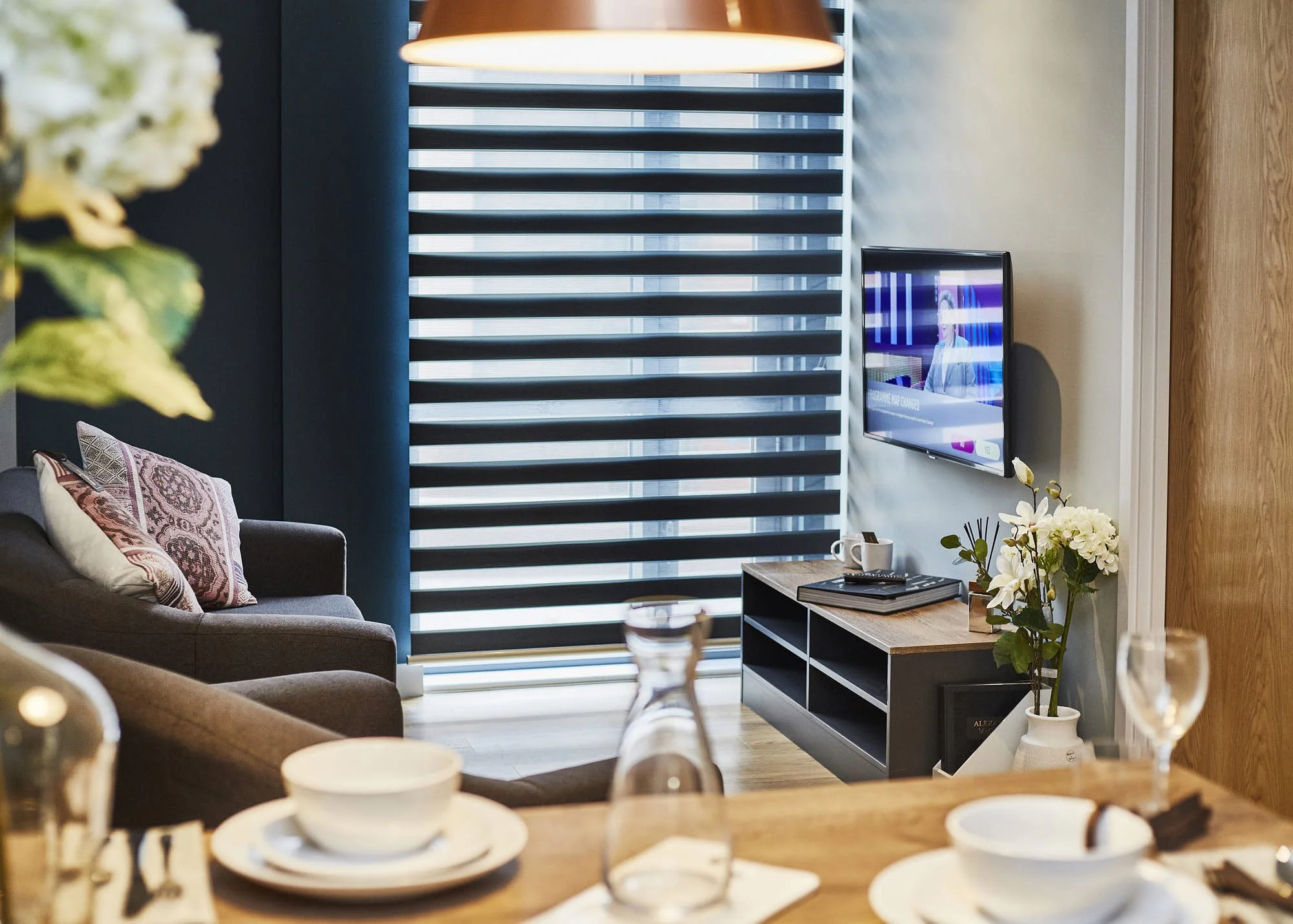 A living room with a dark sofa with patterned pillows, a wooden coffee table with breakfast dishes, a modern media console with a plant, a flat-screen TV, a window with black and white horizontal blinds, a large hanging light, and decorative flowers.