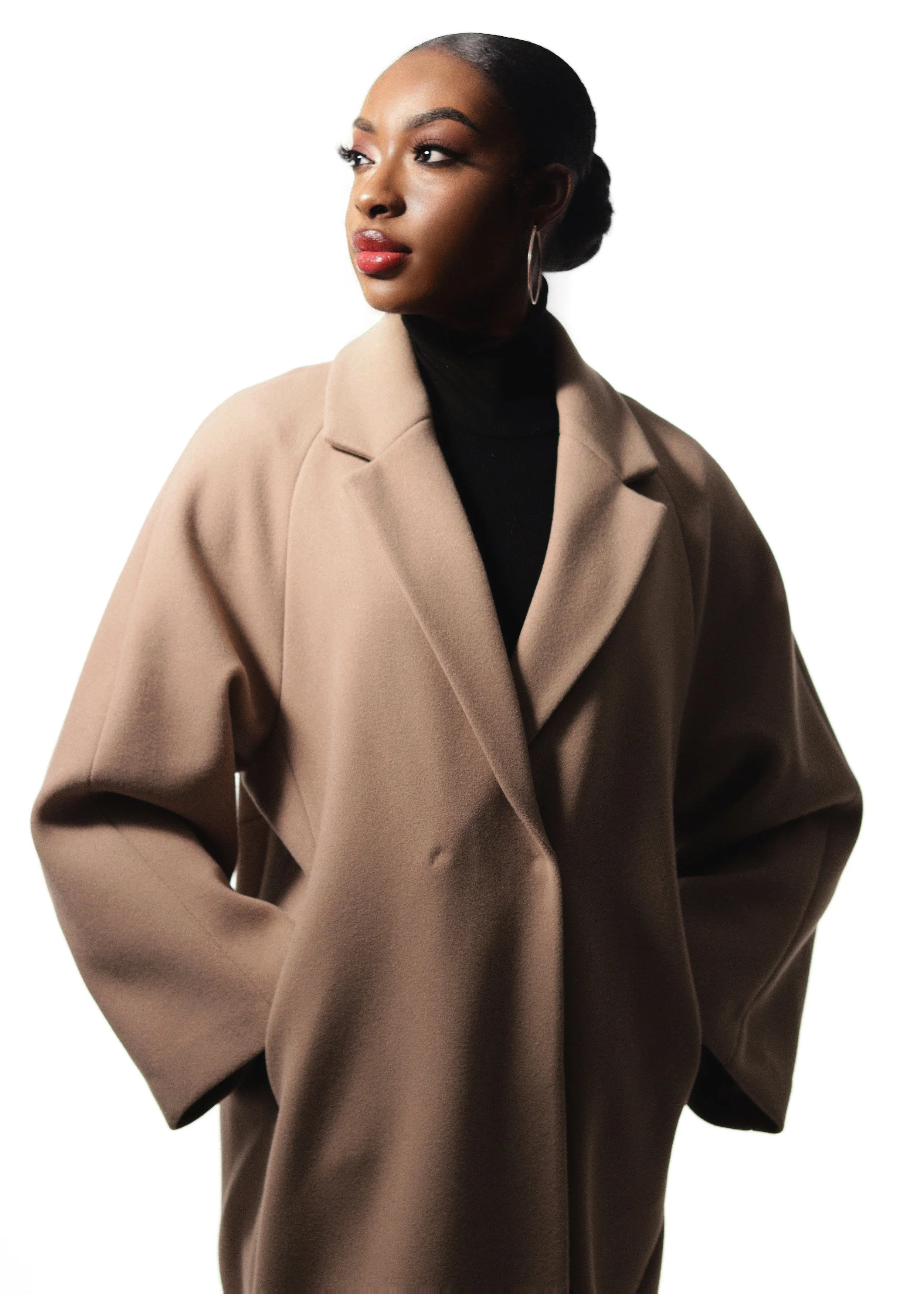 Portrait of a woman with dark skin, pulled-back hair, wearing hoop earrings, a black turtleneck, and a beige coat, looking to her left against a plain white background.