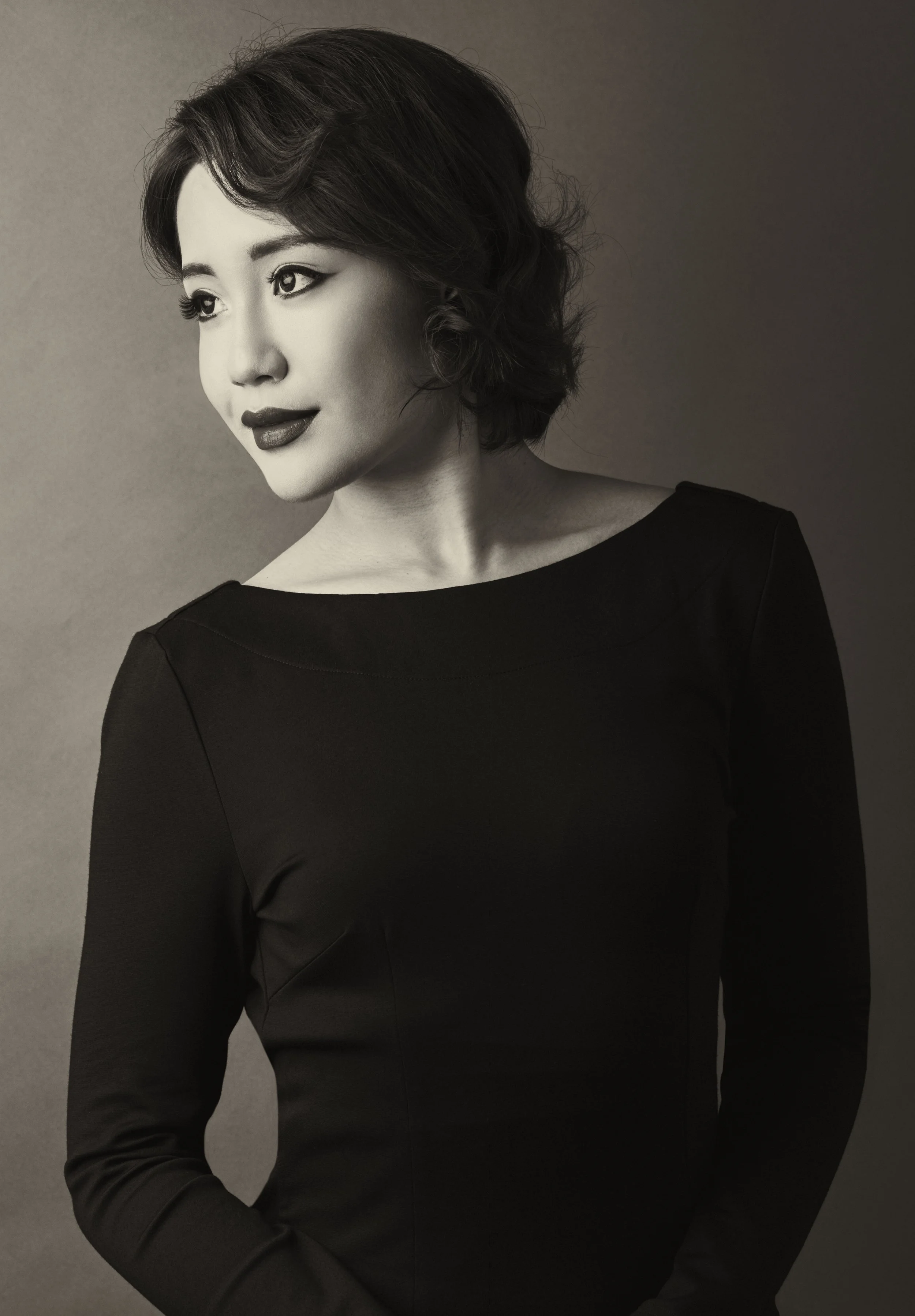 A black-and-white portrait of a woman with short, wavy hair, wearing a dark long-sleeve top, looking to her left with a confident expression.
