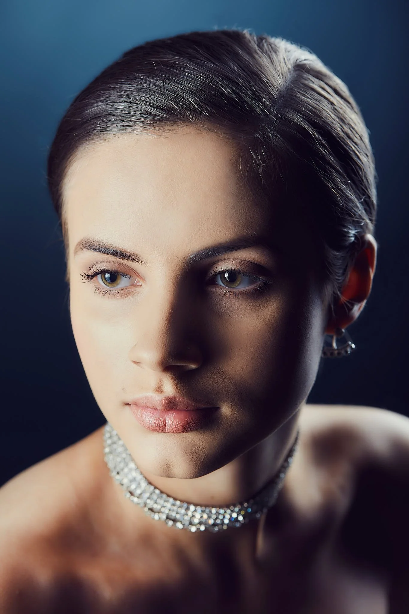 Close-up portrait of a woman with short, dark hair, light skin, wearing a sparkling diamond necklace and earrings, against a dark background.
