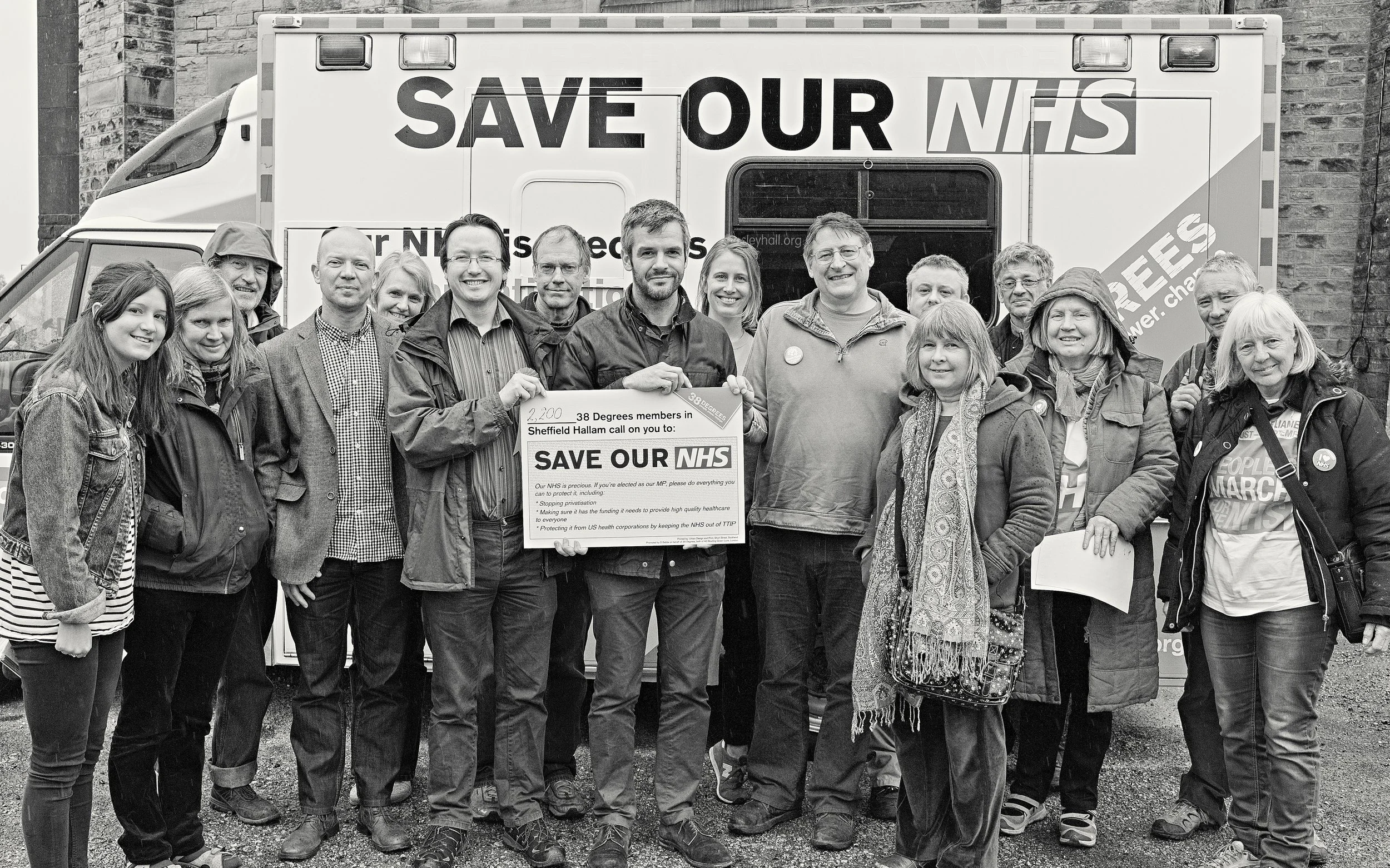Group of diverse people standing together in front of a mobile health unit with a sign that reads "Save Our NHS." They are holding a protest placard calling for preserving the UK’s National Health Service, showing support for healthcare funding.