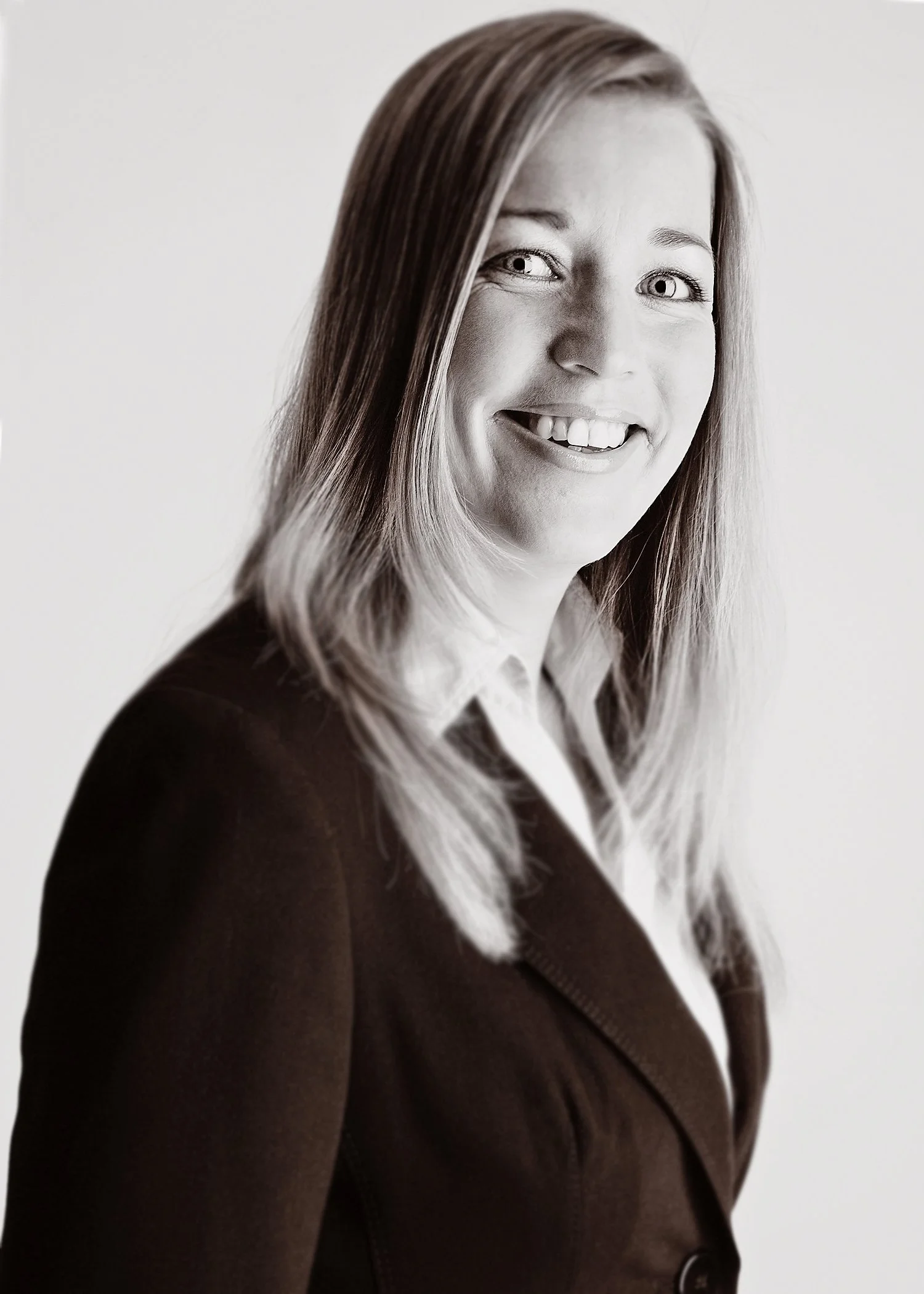 Black and white portrait of a smiling woman with long hair, wearing a dark blazer and white shirt, against a light background.