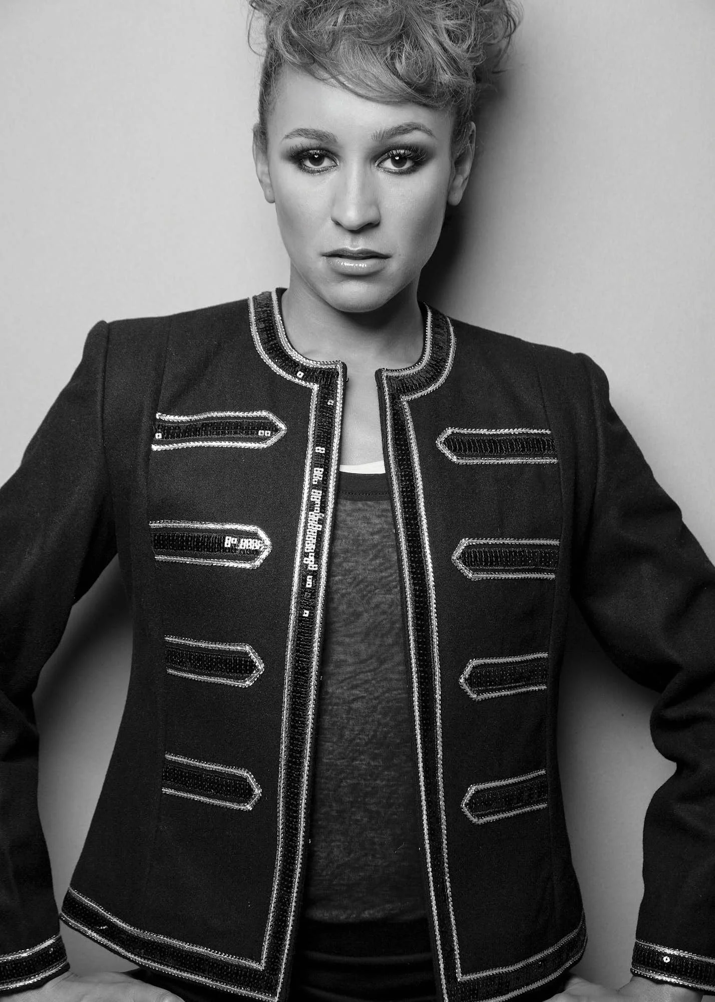 Black and white photo of a woman with short, curly hair wearing a dark jacket with metallic embellishments and a light-colored shirt underneath, posing against a plain wall.
