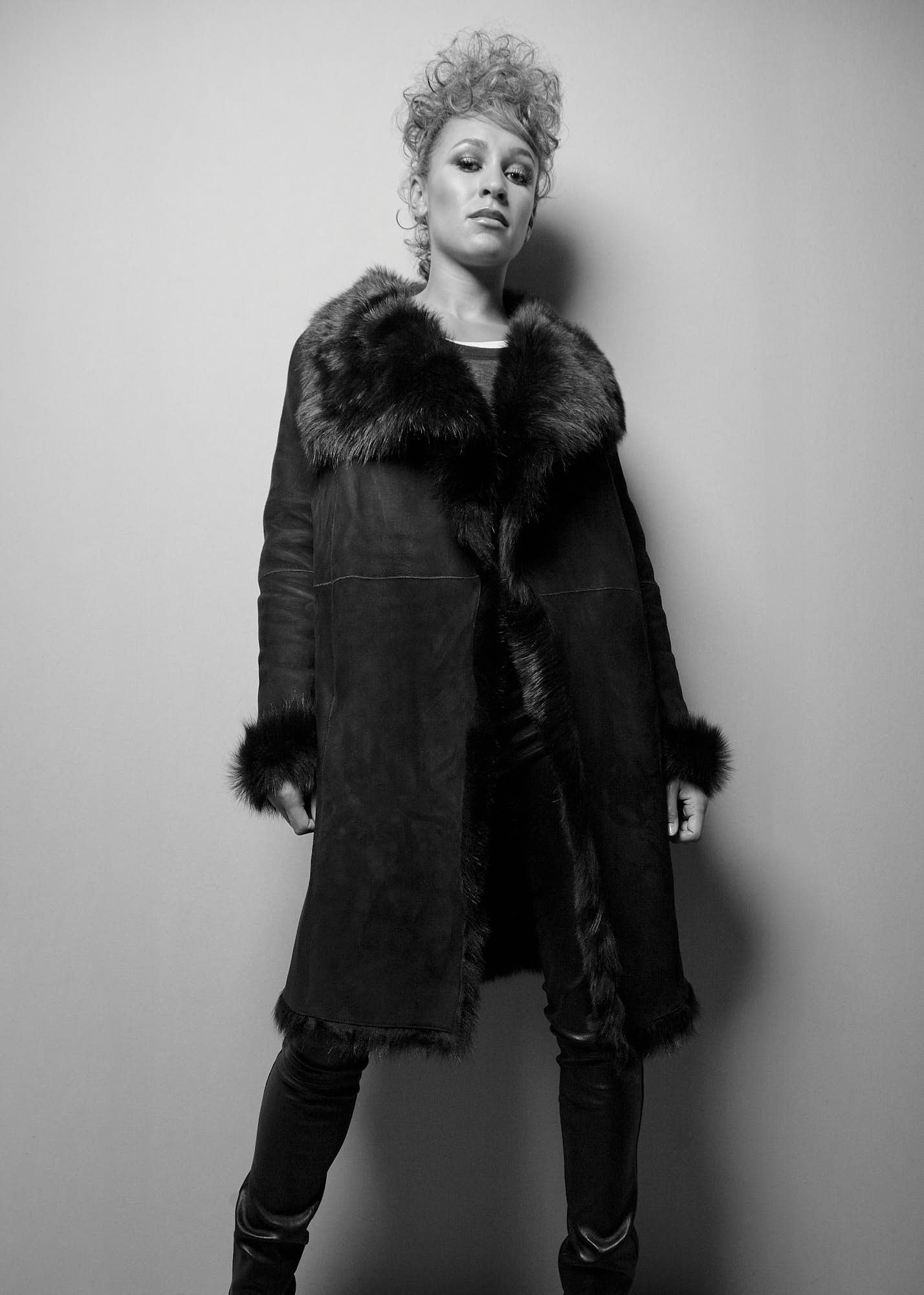 Black and white photo of a woman with curly hair standing against a plain wall, wearing a long coat with a fur collar and fur trim on the sleeves and down the front.