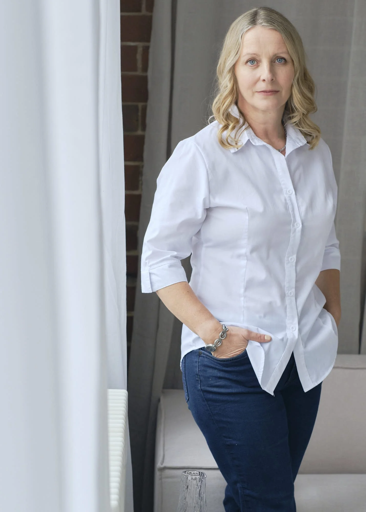 A woman with blonde hair wearing a white button-down shirt and blue jeans standing indoors, with a brick wall and a curtain behind her, and a glass on the floor in front of her.