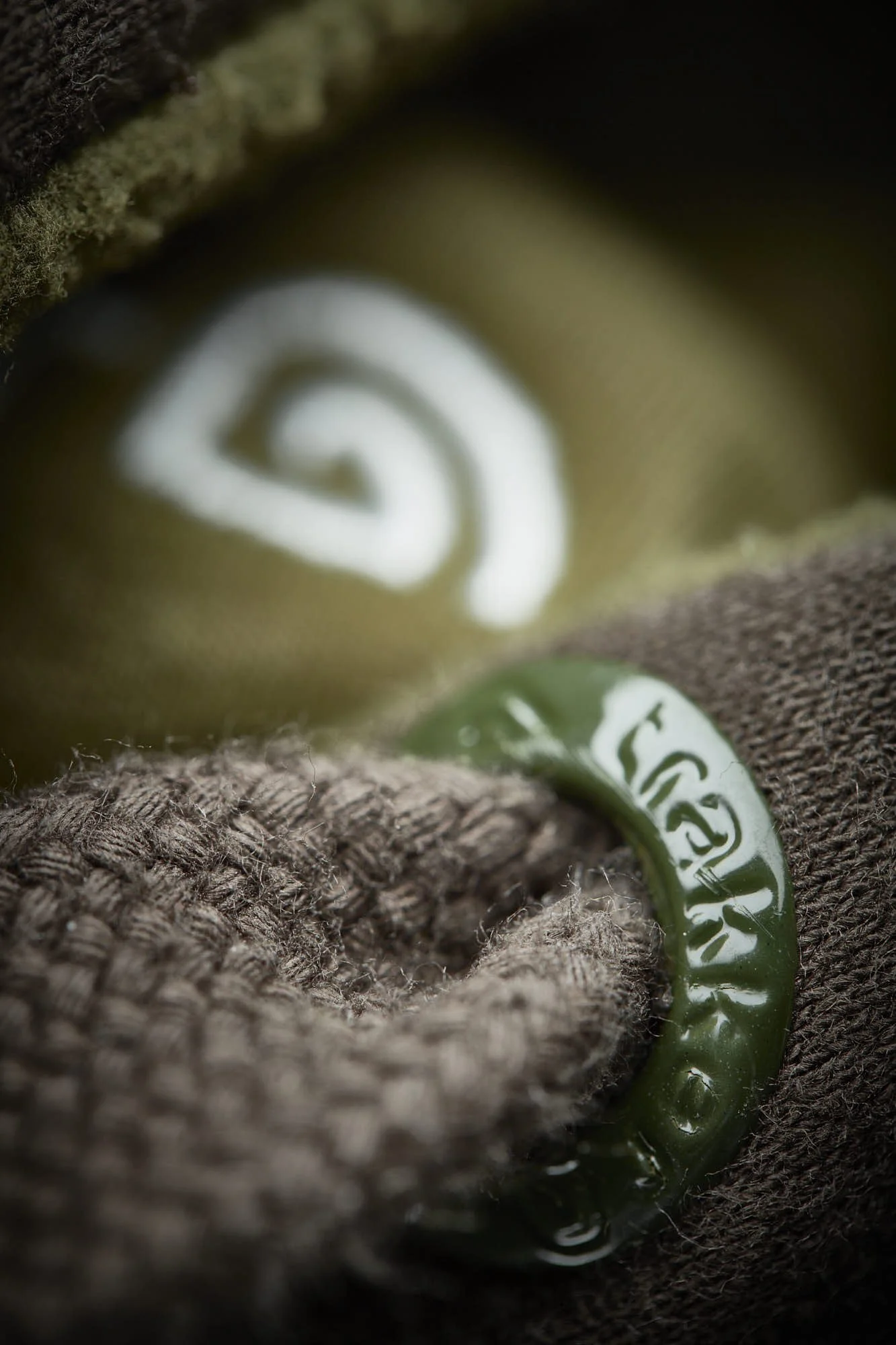 A close-up photograph of a green and white moldavite teardrop-shaped pendant with a swirled design on a rough, textured brown fabric surface.