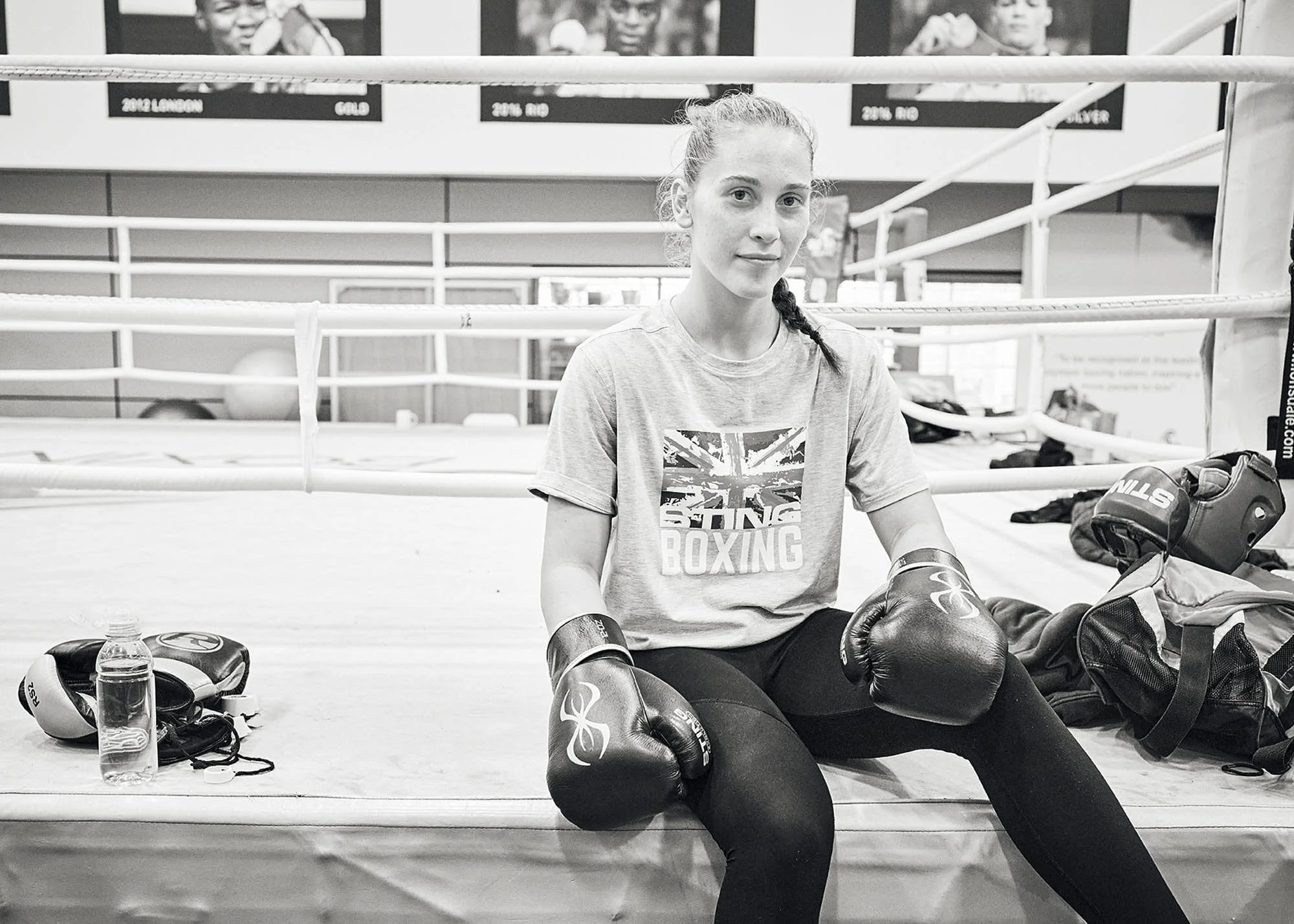 A young woman sitting inside a boxing gym, wearing boxing gloves, with boxing equipment and a water bottle nearby. The gym has boxing rings and motivational posters on the wall.