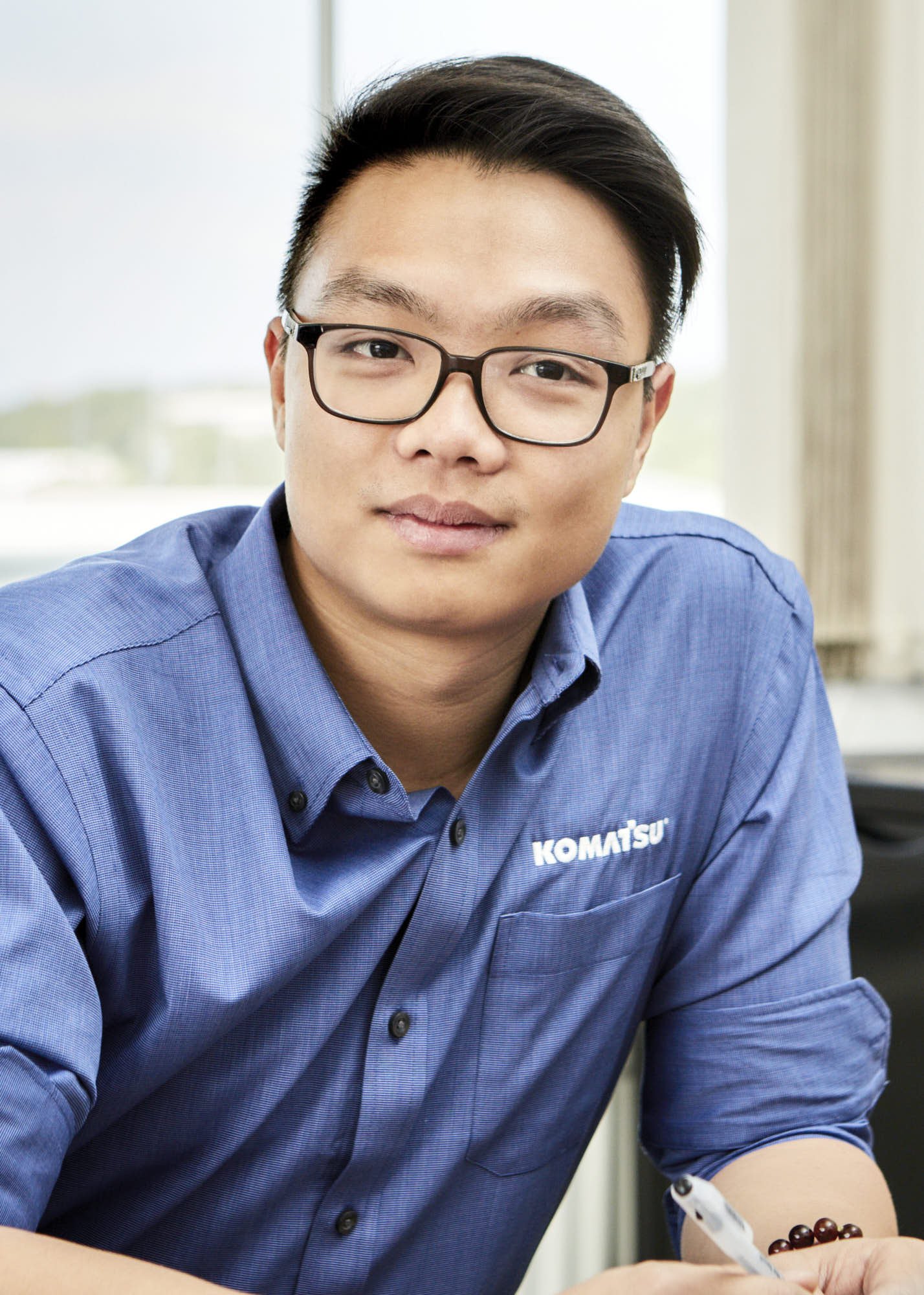A young man with short black hair and glasses wearing a blue Komatsu shirt, sitting indoors with a white background, holding a pen, and wearing a bracelet on his left wrist.