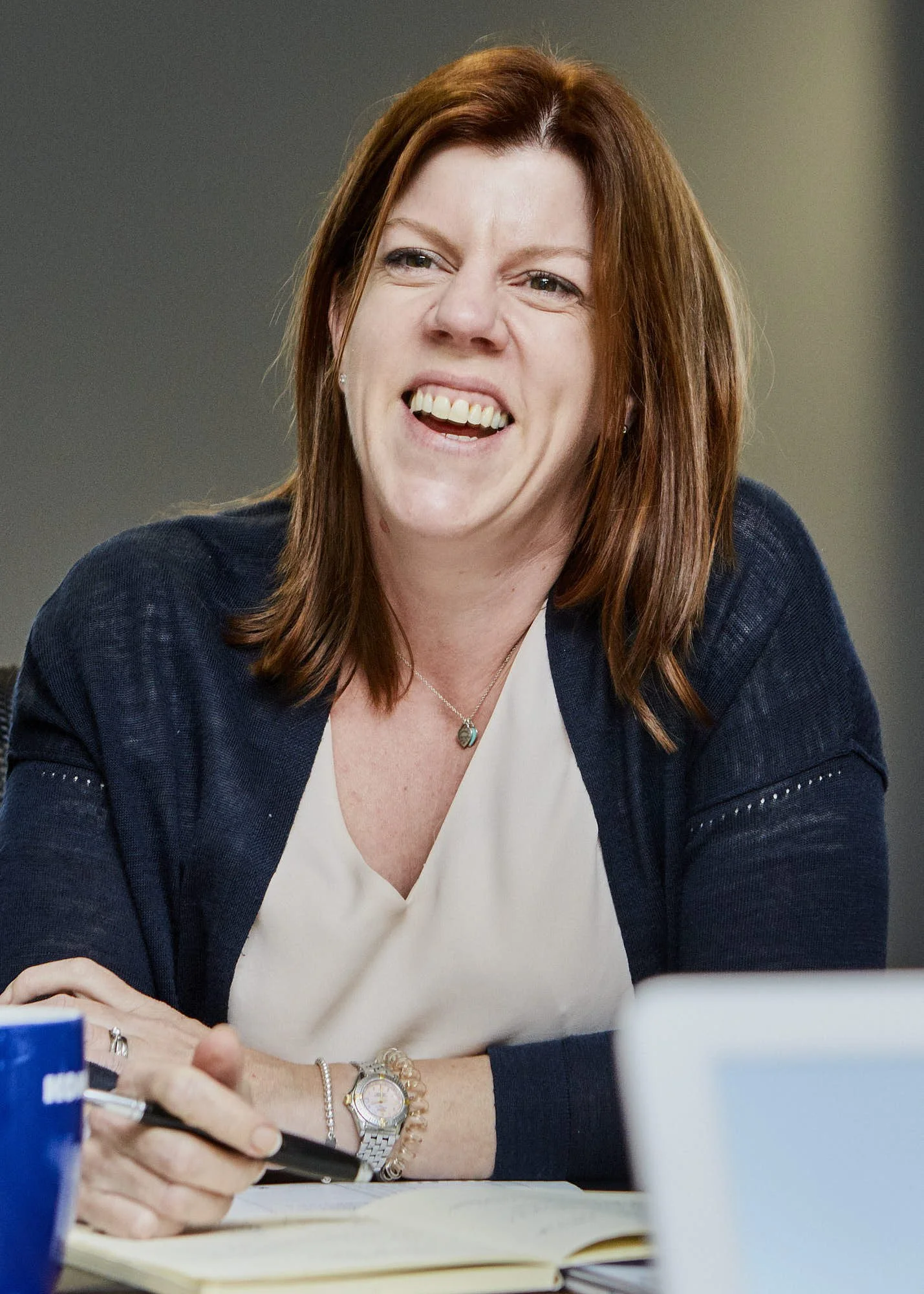 A woman with red hair smiling and looking off to the side, seated at a table with papers, a pen, and a blue notebook in front of her.