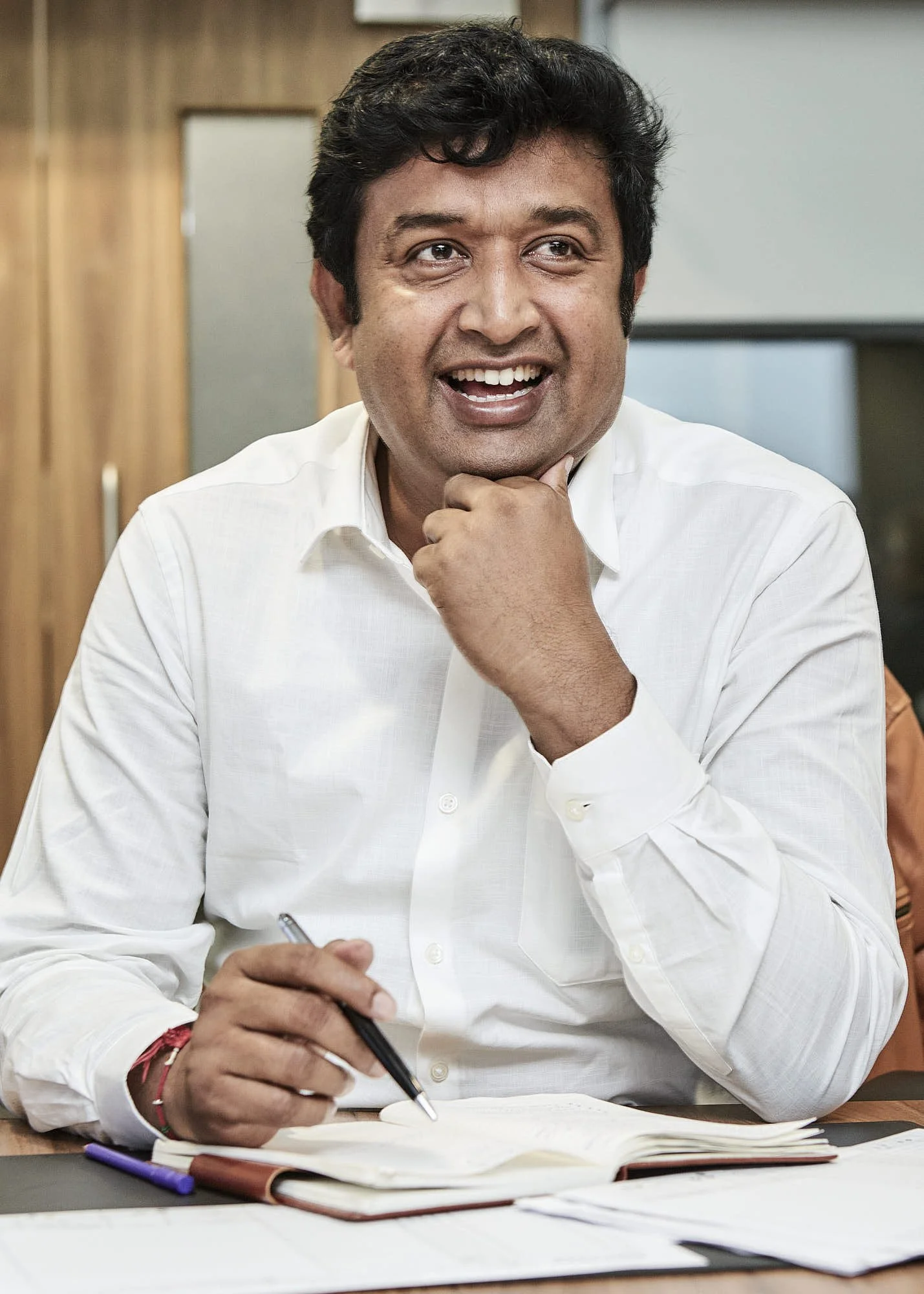 A man wearing a white shirt is smiling and sitting at a desk. He has dark curly hair, and is holding a pen while looking to his right. There are papers and writing instruments on the desk.