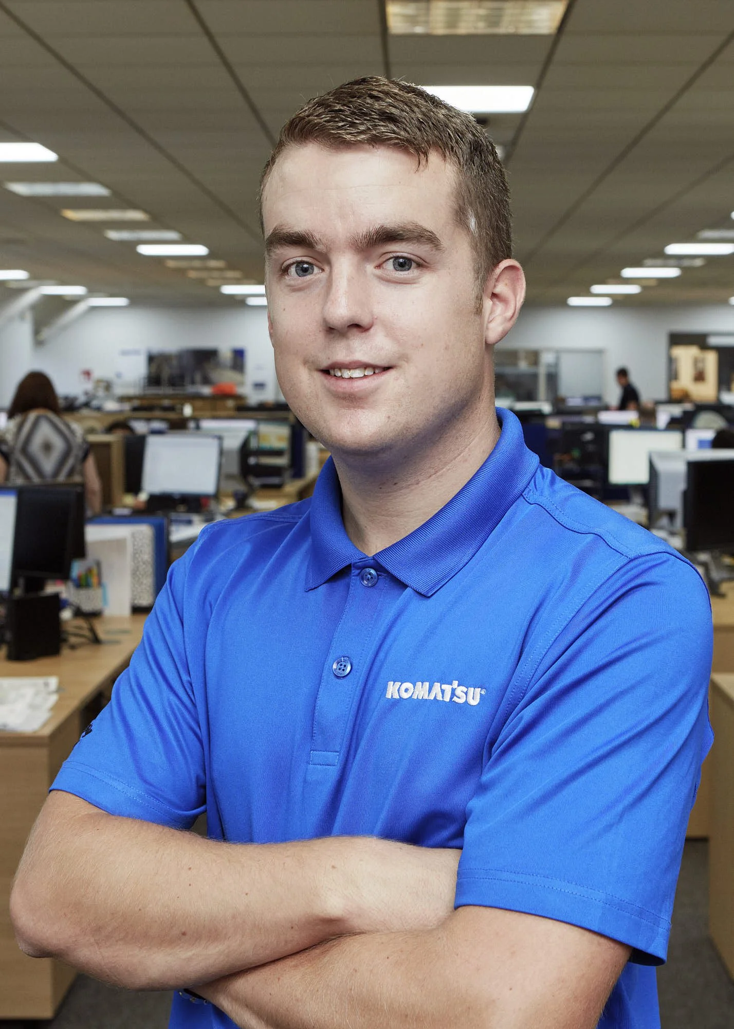 A man in a blue Komatsu uniform is standing with crossed arms in an office environment with multiple computer workstations in the background.