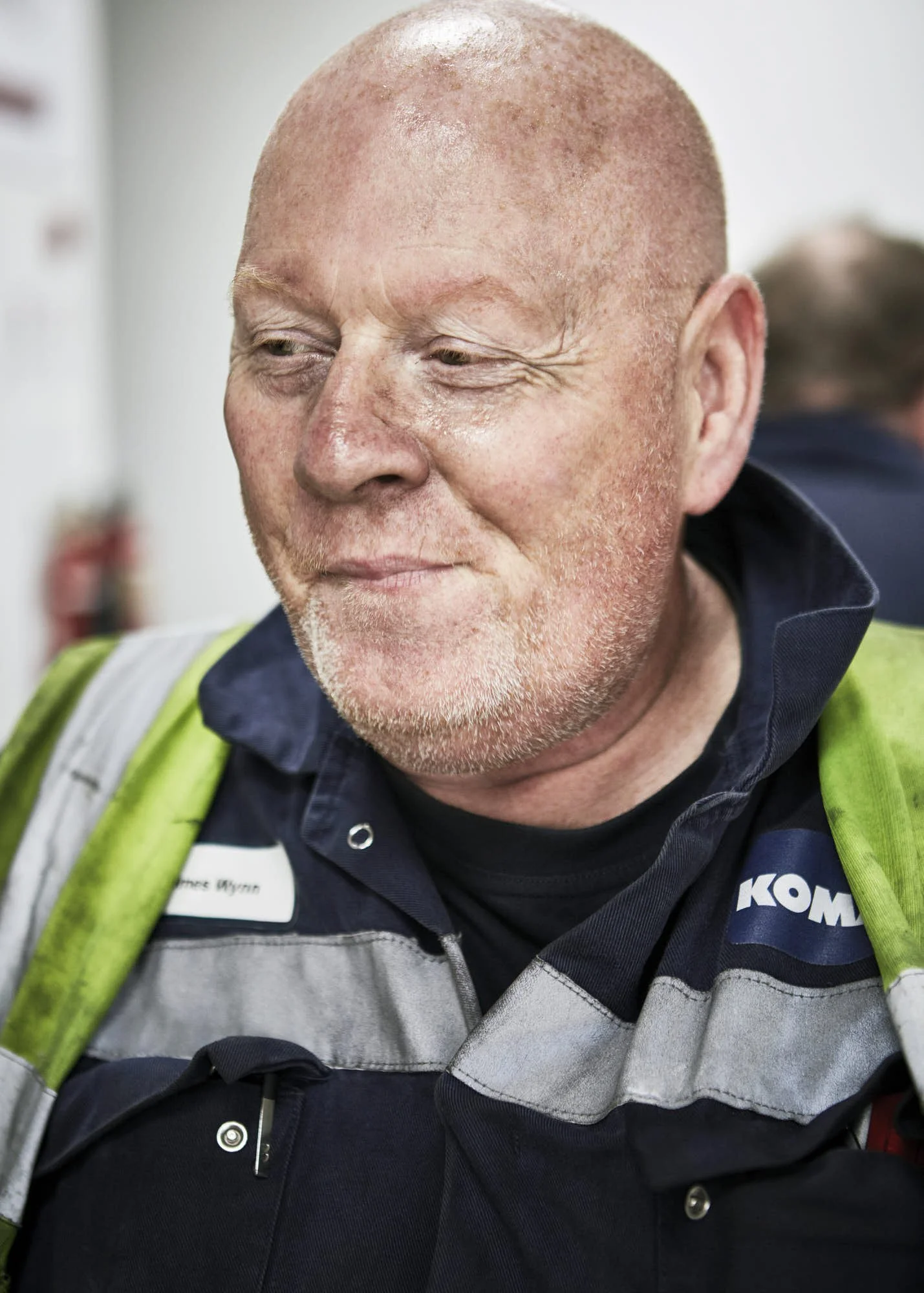 Close-up of a smiling man with a shaved head, wearing a uniform with a reflective vest and a name tag, in an indoor setting.