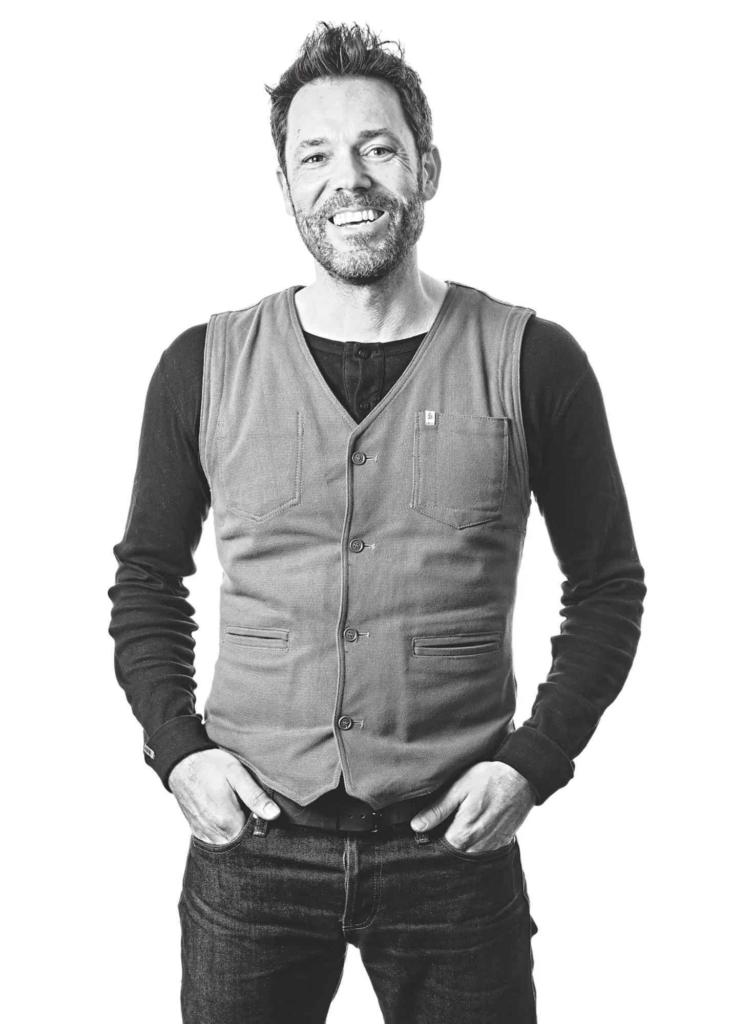 Black and white portrait of a man smiling, wearing a vest over a long-sleeve shirt, standing against a plain white background.