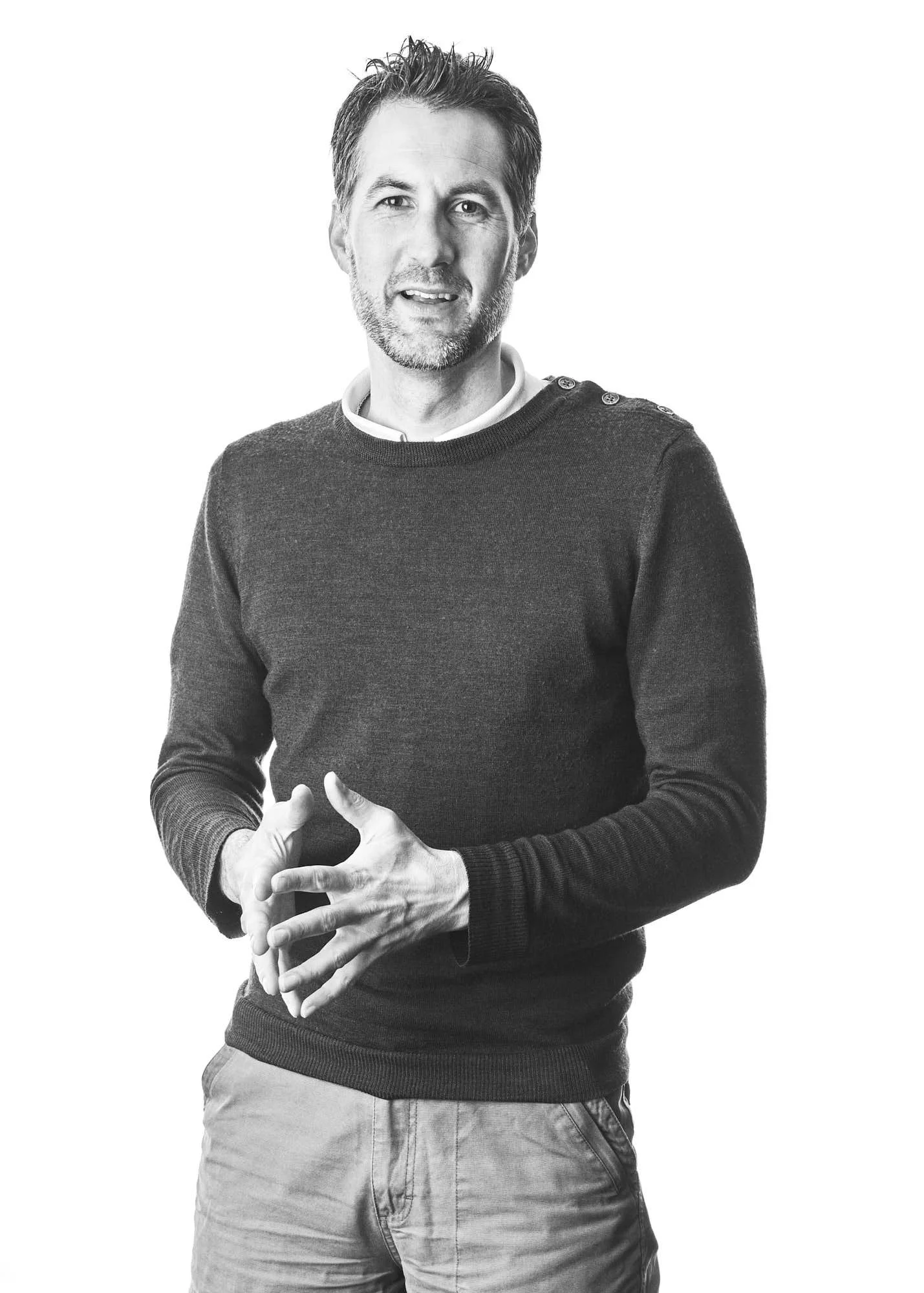 A black and white photo of a middle-aged man with short hair and a beard, wearing a dark sweater with button details on the shoulder and light-colored pants, standing against a plain background.