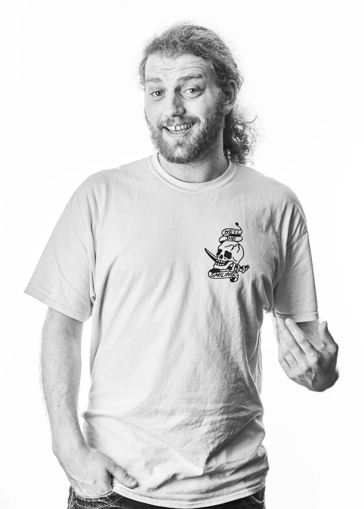 Black and white photo of a man with long hair, a beard, wearing a t-shirt with a skull and text design, smiling with his teeth showing, pointing to his side with his right hand.
