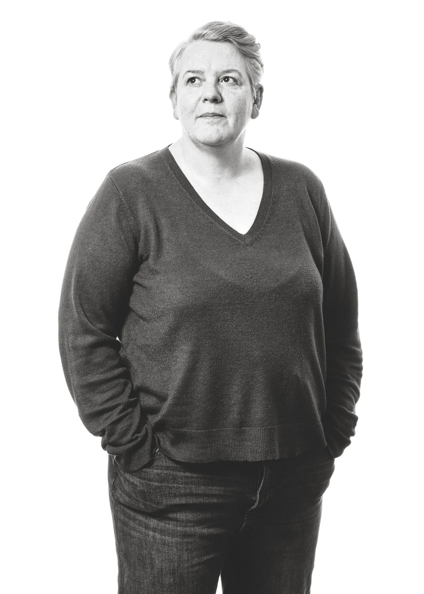 Black and white photo of a woman with short hair wearing a V-neck sweater and dark jeans, standing with one hand in her pocket against a plain white background.
