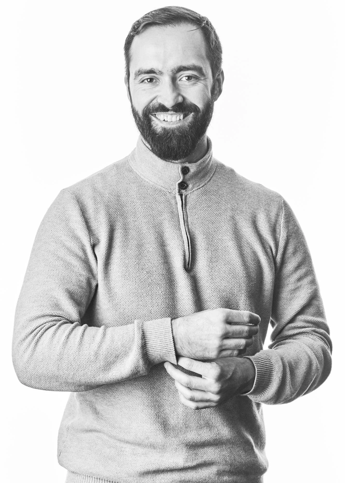 Black and white portrait of a man with a beard, smiling, wearing a zip-up sweater with a high collar.