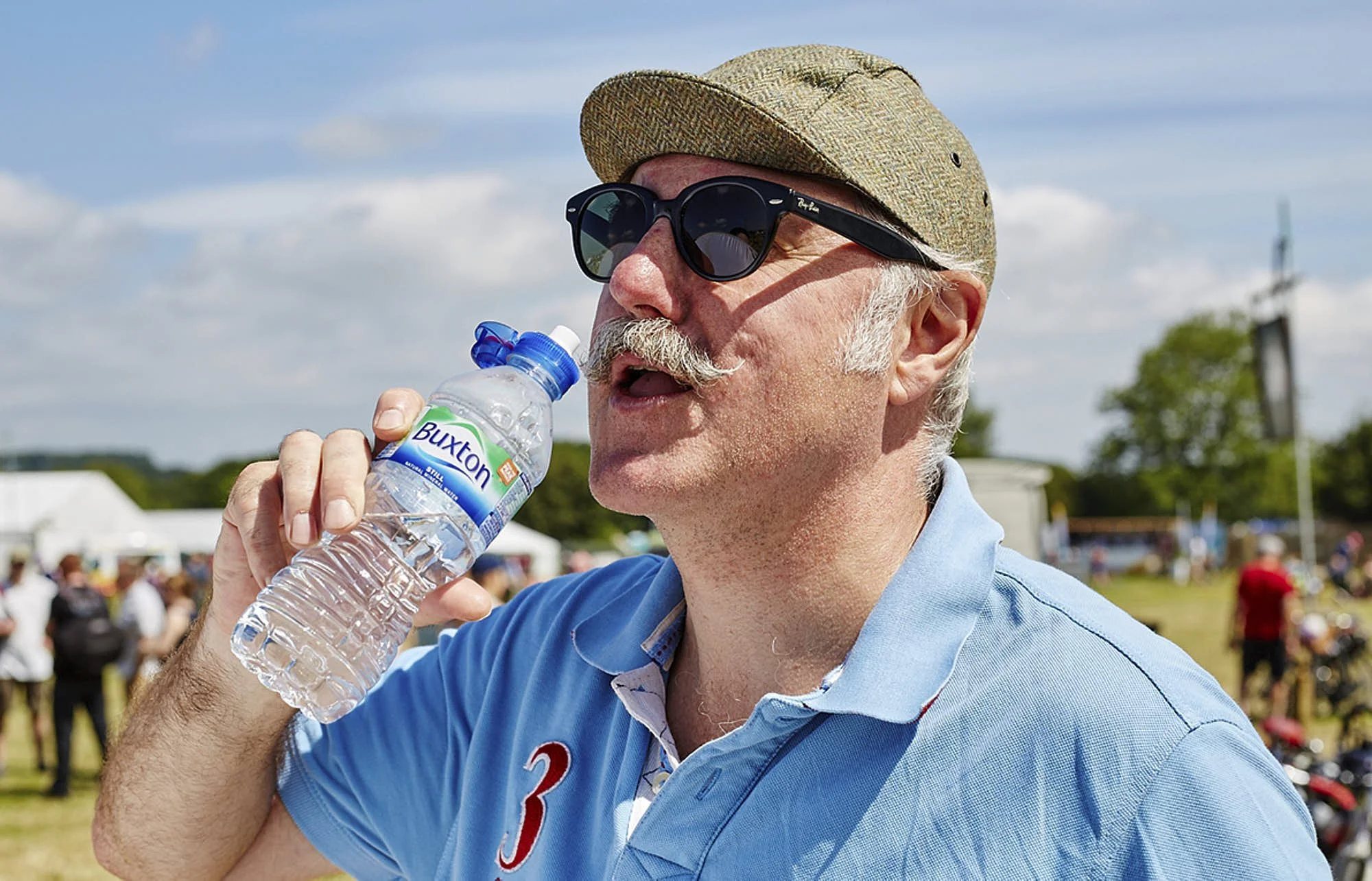 A man wearing sunglasses, a beige cap, and a light blue polo shirt is drinking water from a plastic bottle at an outdoor event, with a crowd and cloudy sky in the background.