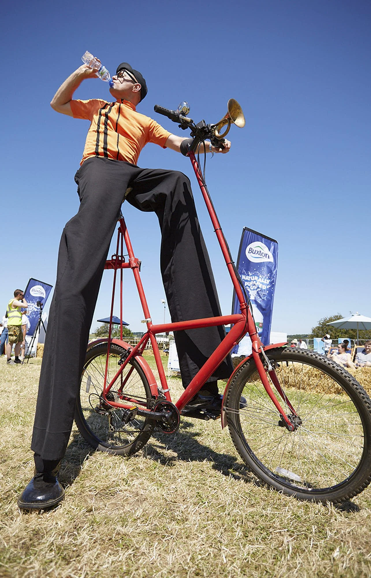 A person standing on a small red bicycle, drinking water from a plastic bottle, wearing an orange shirt, black pants, and a cap, outdoors on a sunny day.