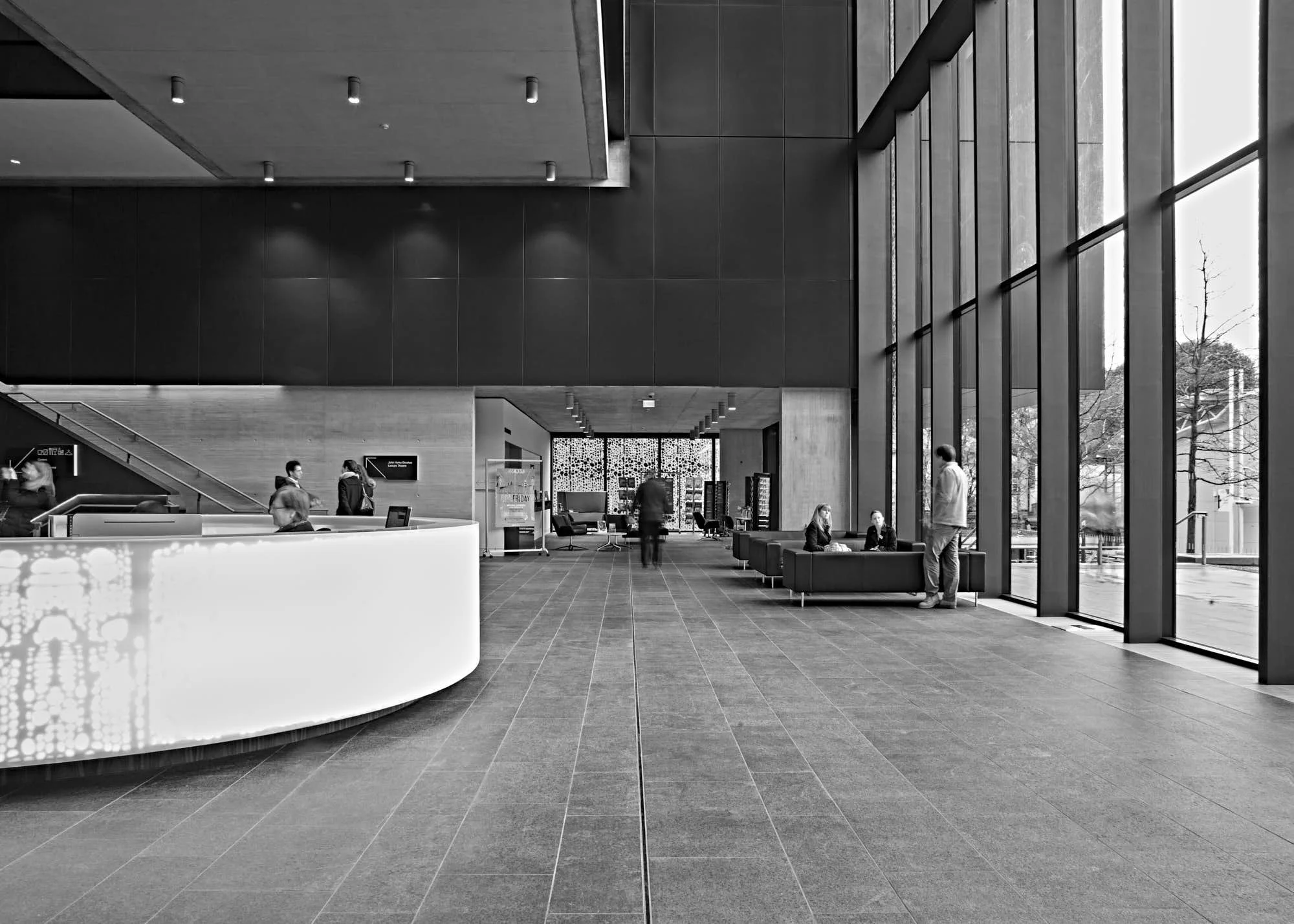 Modern building lobby with high glass windows, seating area with black sofas, people walking and sitting, reception desk in the foreground, and an escalator on the left.