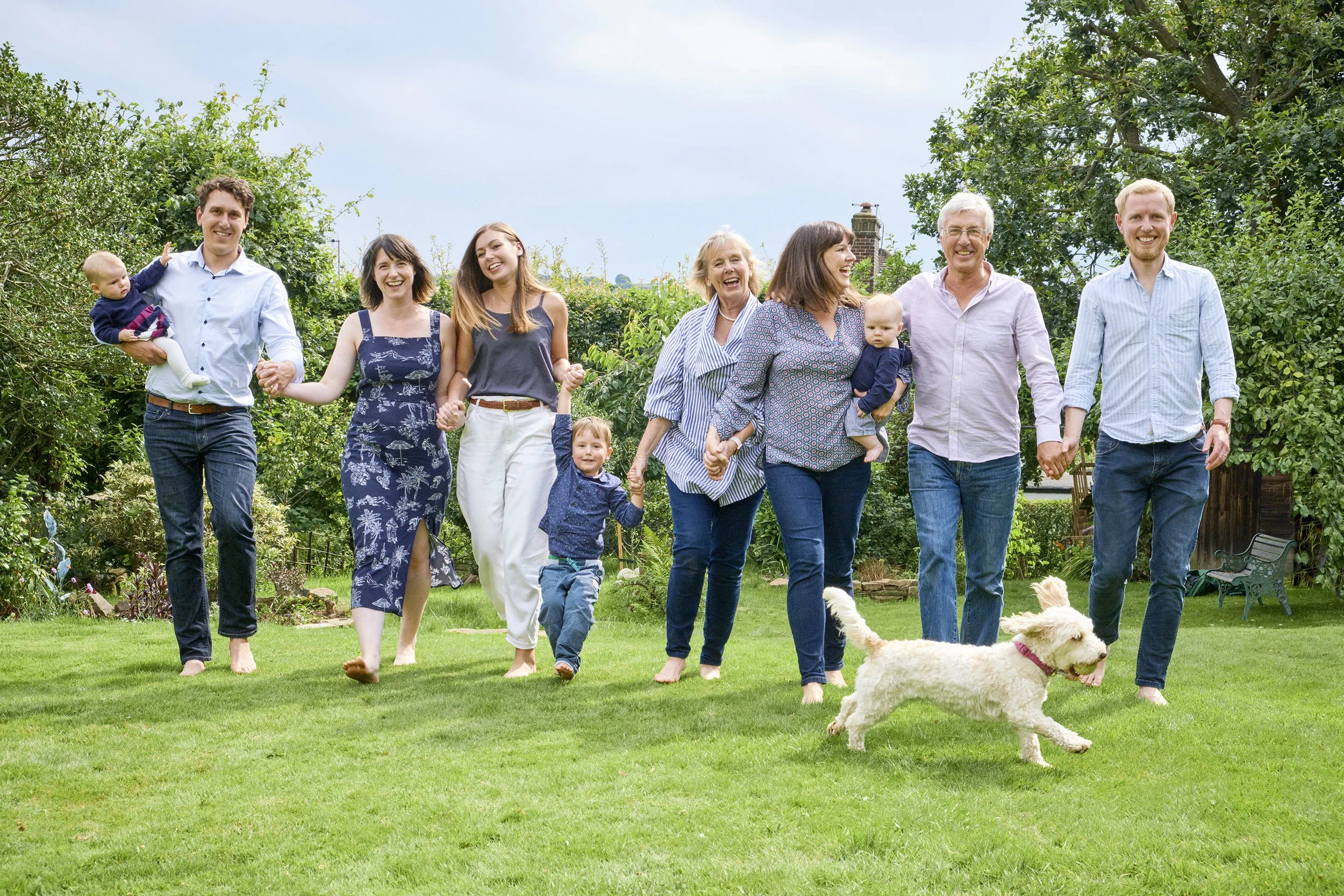 A group of nine people, including children and adults, are walking together in a lush green garden, holding hands and smiling. There are dogs in the scene, one being led by a woman. The group appears joyful and relaxed, enjoying time outdoors on a br