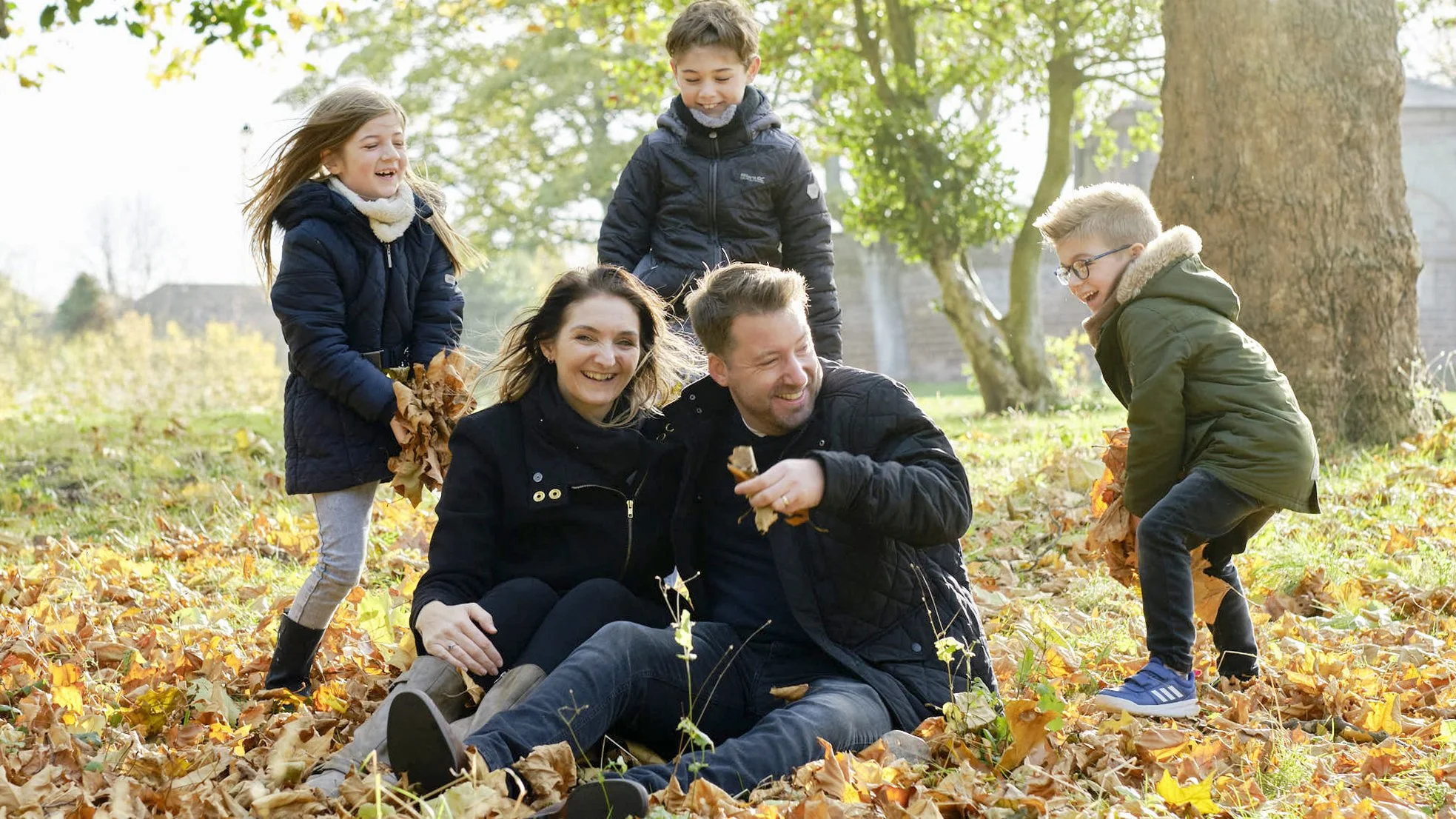 Family of five enjoying a fall day outdoors among fallen leaves, with children playing and parents sitting on the ground smiling.