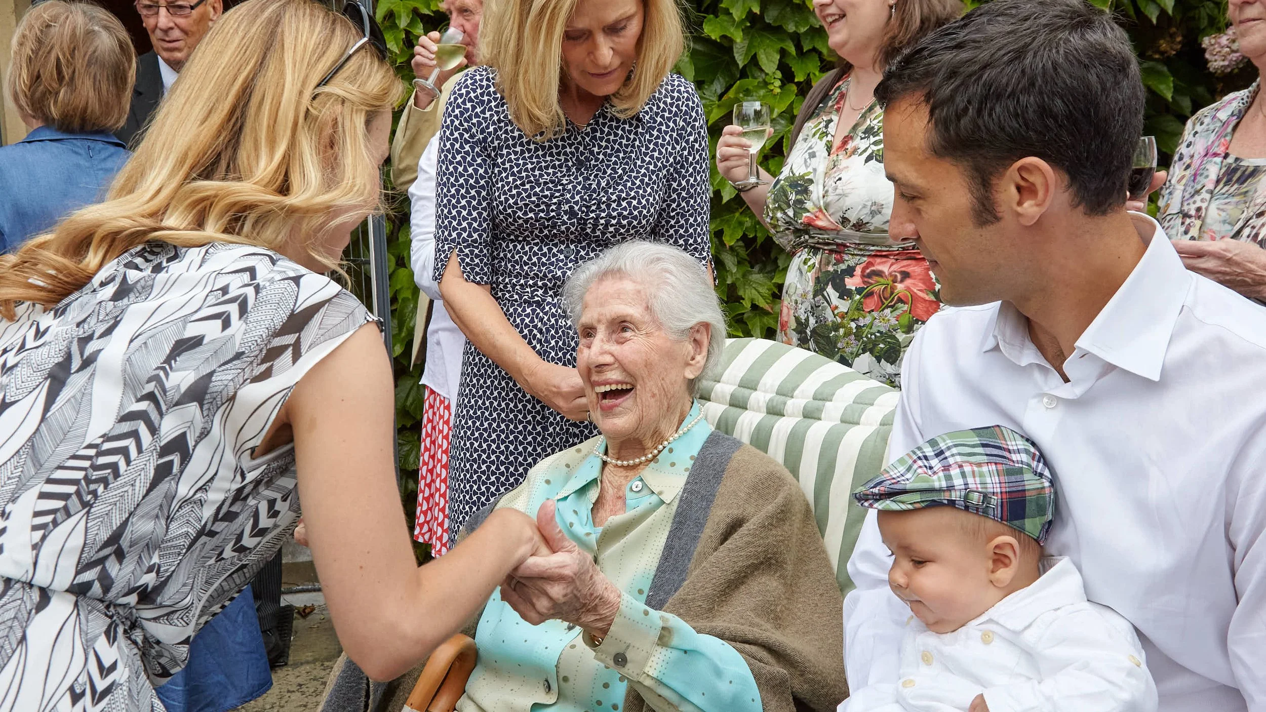 A group of people, including an elderly woman seated in a chair, are gathered outdoors. They are smiling and engaging in conversation, with some holding glasses of wine. The elderly woman is shaking hands and smiling at a younger woman with long blon