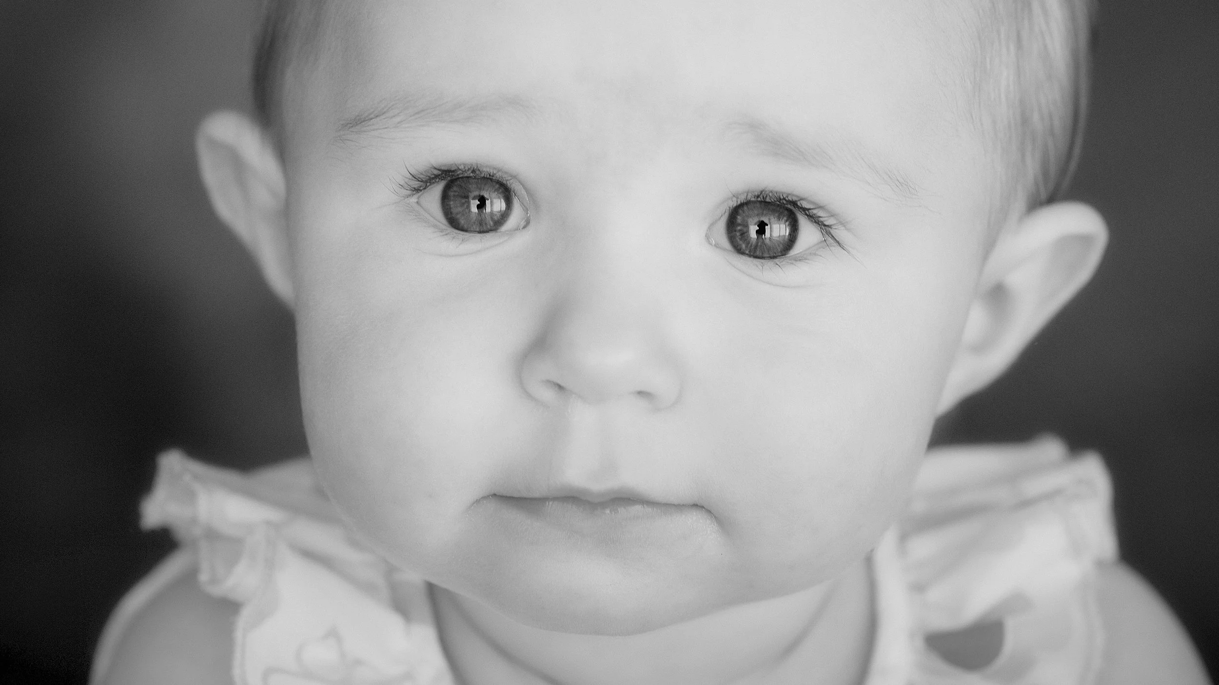 Close-up black and white photo of a child's face, focusing on large eyes reflecting a person and a window, with soft lighting highlighting facial features.