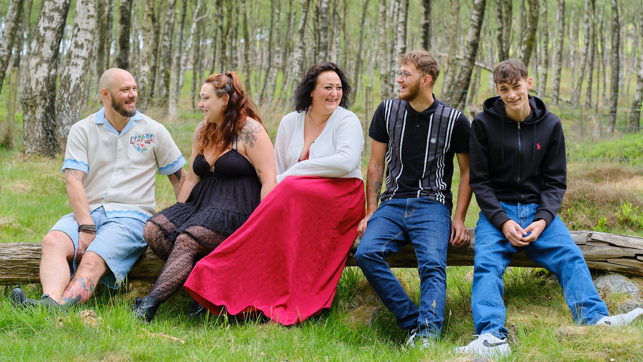 Group of five people sitting on a log outdoors in a forest, smiling and engaging with each other.