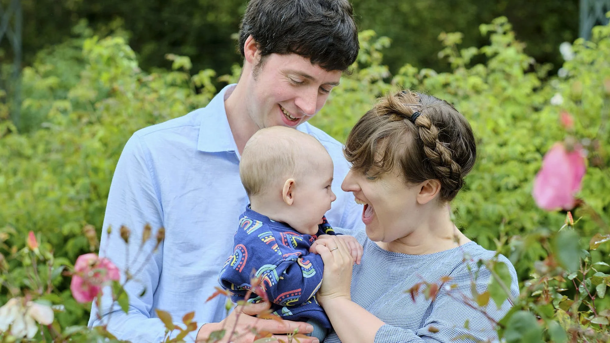A smiling family of four, including a father, mother, and two young children, sharing joyful moments outdoors among flowering bushes.