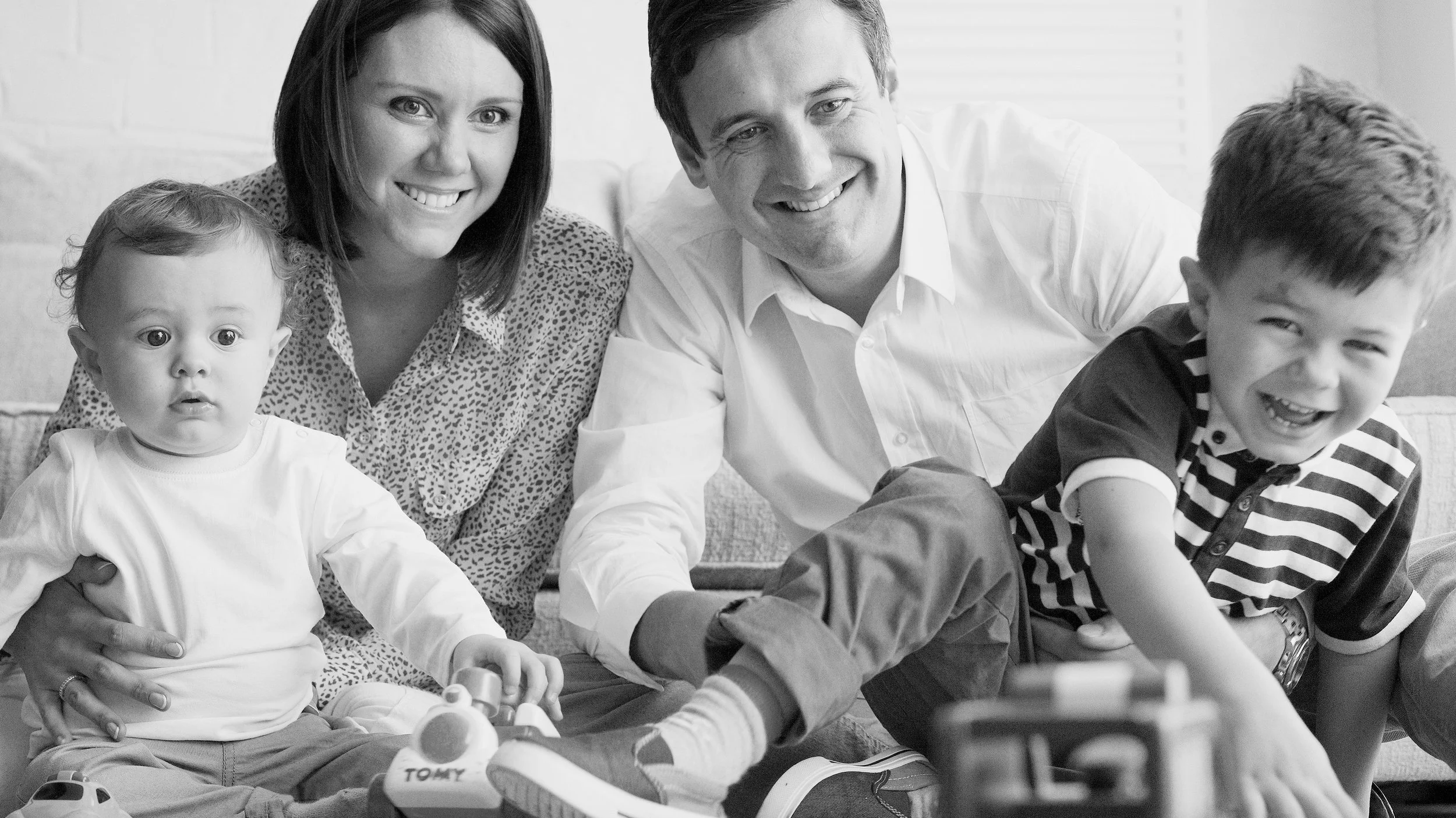 A black-and-white photograph of a happy family of four sitting on a couch and playing with toy cars. The mother, with dark hair and a patterned blouse, is holding a young child with light hair and a white shirt. The father, wearing a light-colored sh