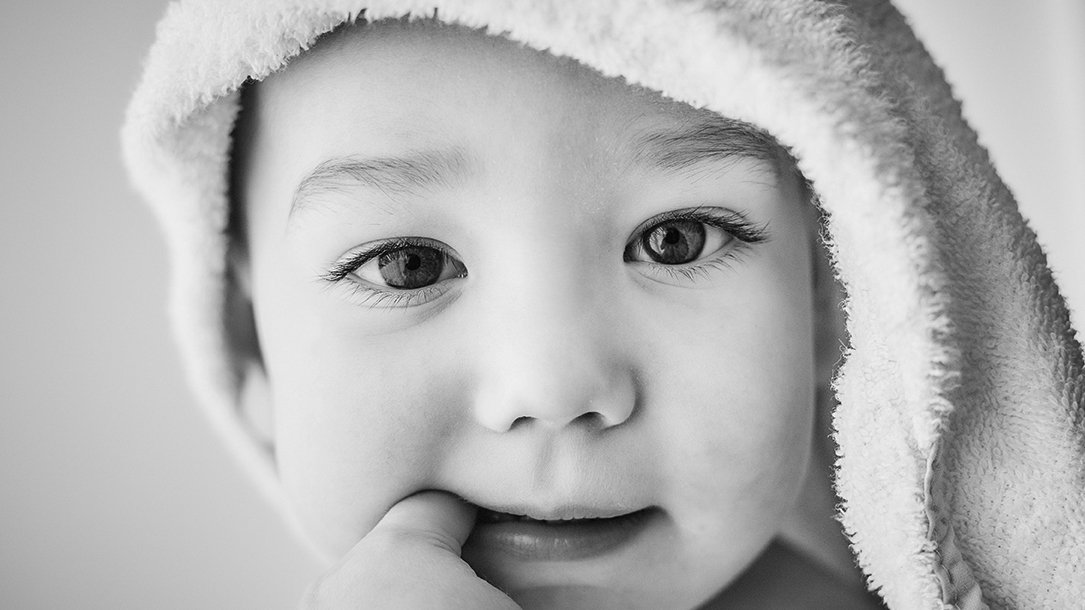 Close-up of a young child with a towel on their head, touching their lips and looking at the camera.